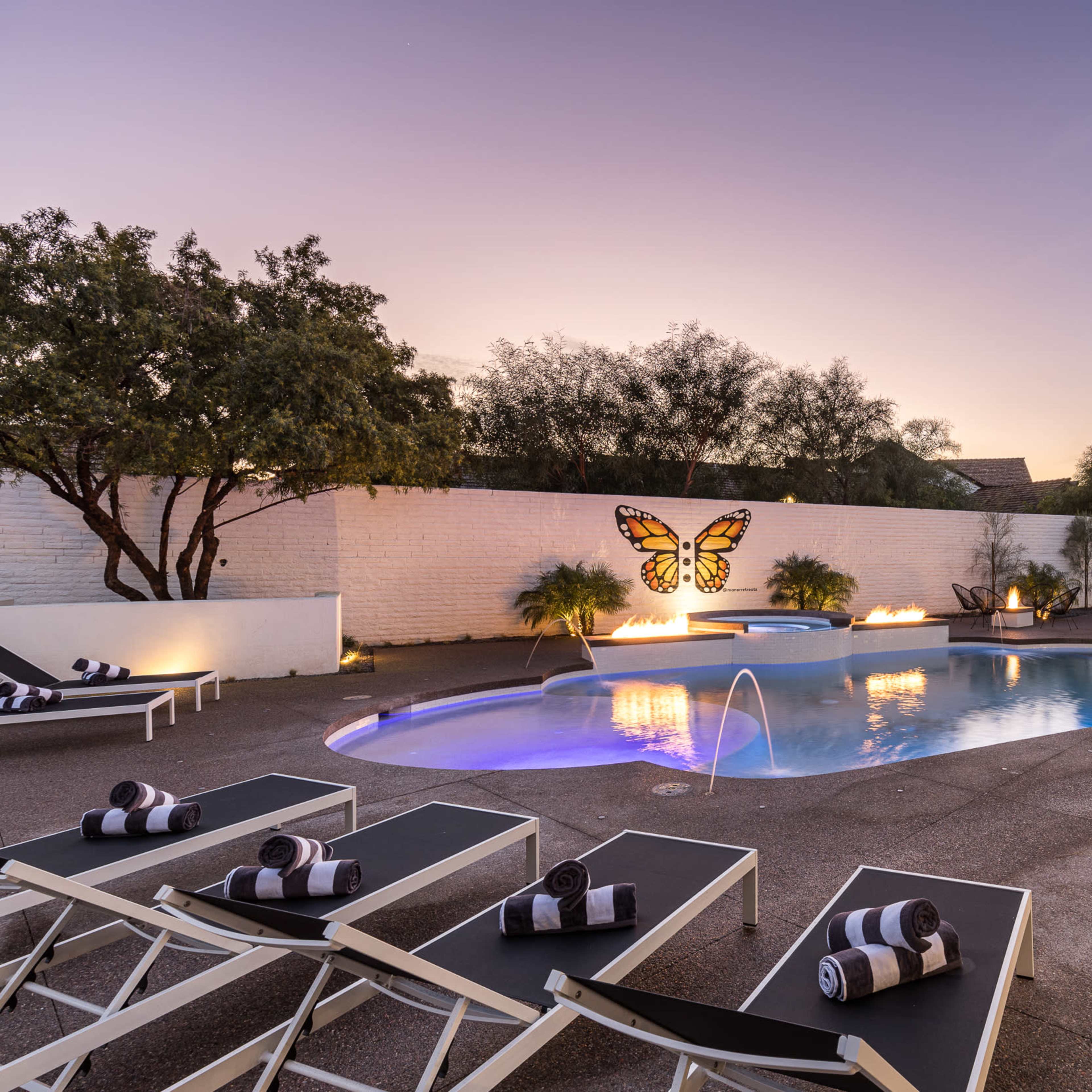 A pool area features a butterfly mural on a wall, surrounded by lounge chairs and lit by flames on either side at dusk.