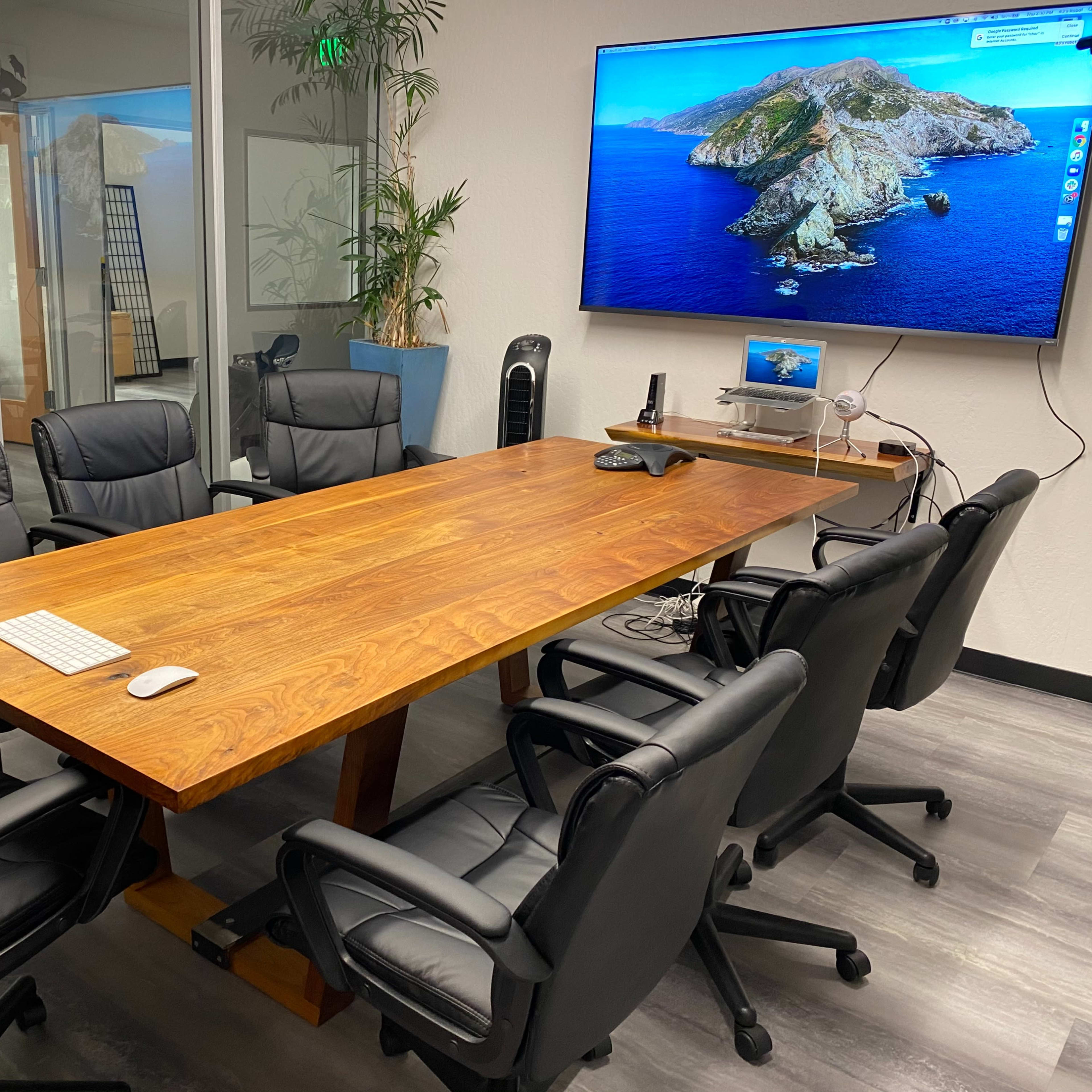 A meeting room features a large wooden table surrounded by six black office chairs, with a wall-mounted screen displaying a coastal image.