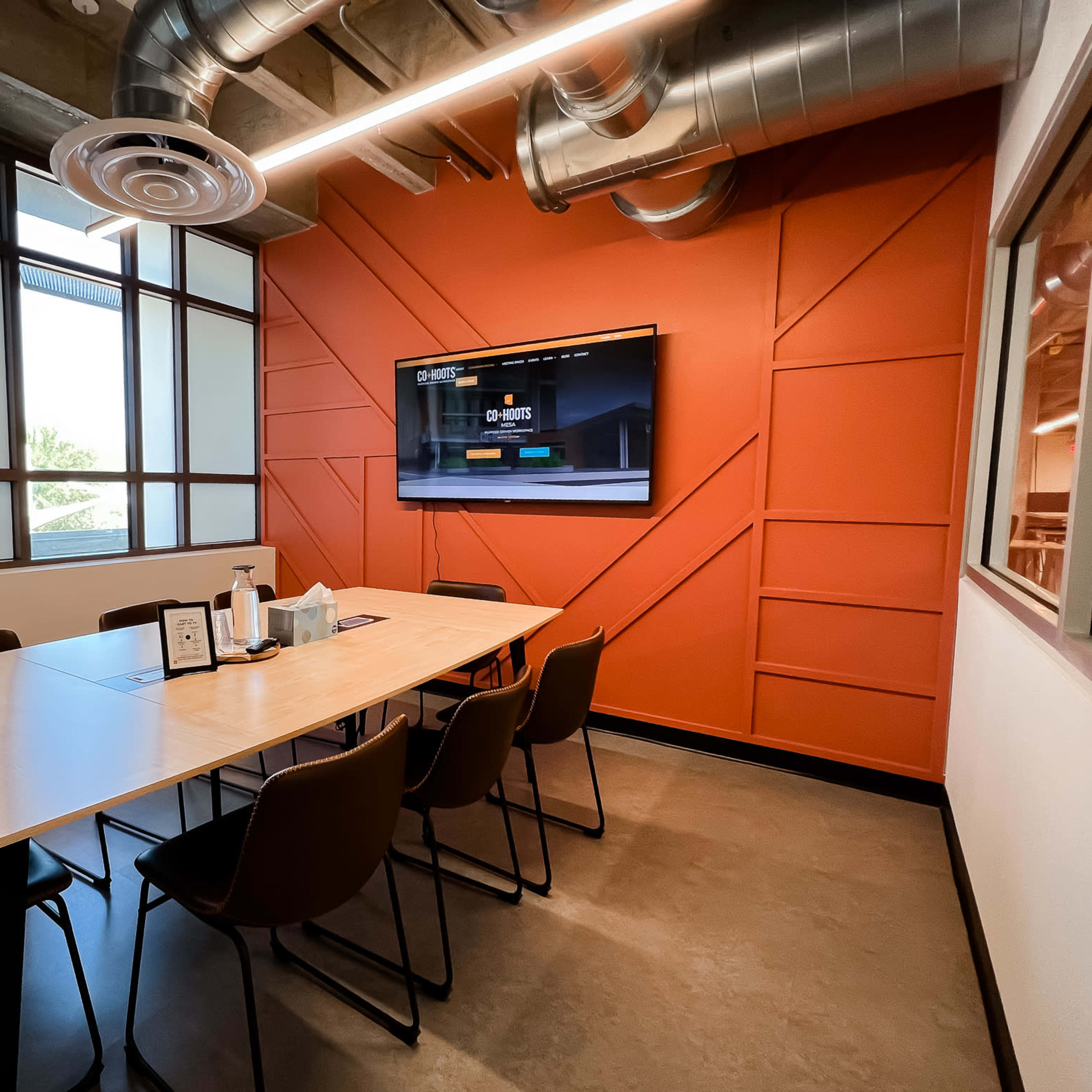 A conference room features a large table surrounded by chairs, with a television mounted on an orange accent wall and large windows providing natural light.