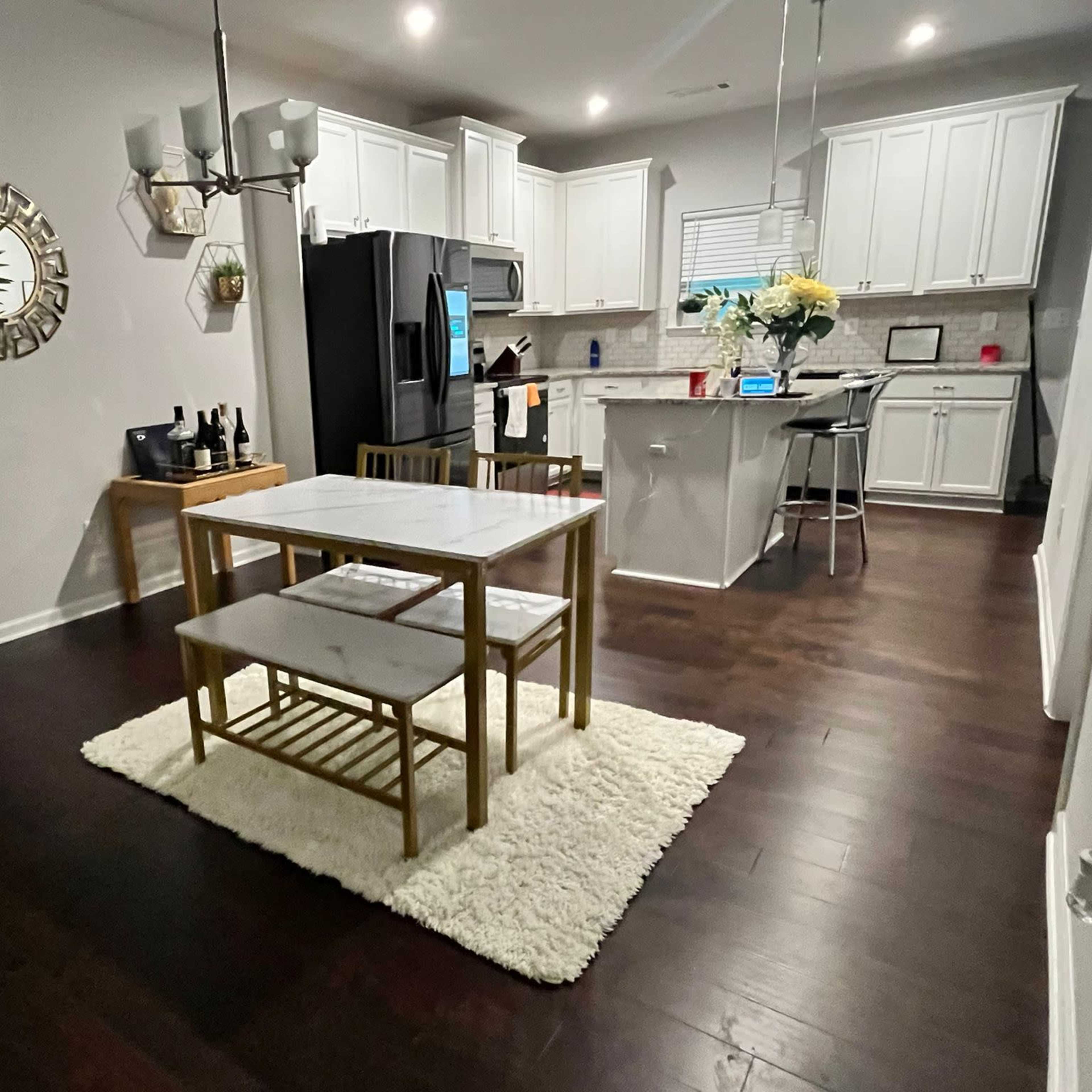 A modern kitchen and dining area features a white table with two benches on a rug, surrounded by cabinetry and appliances.