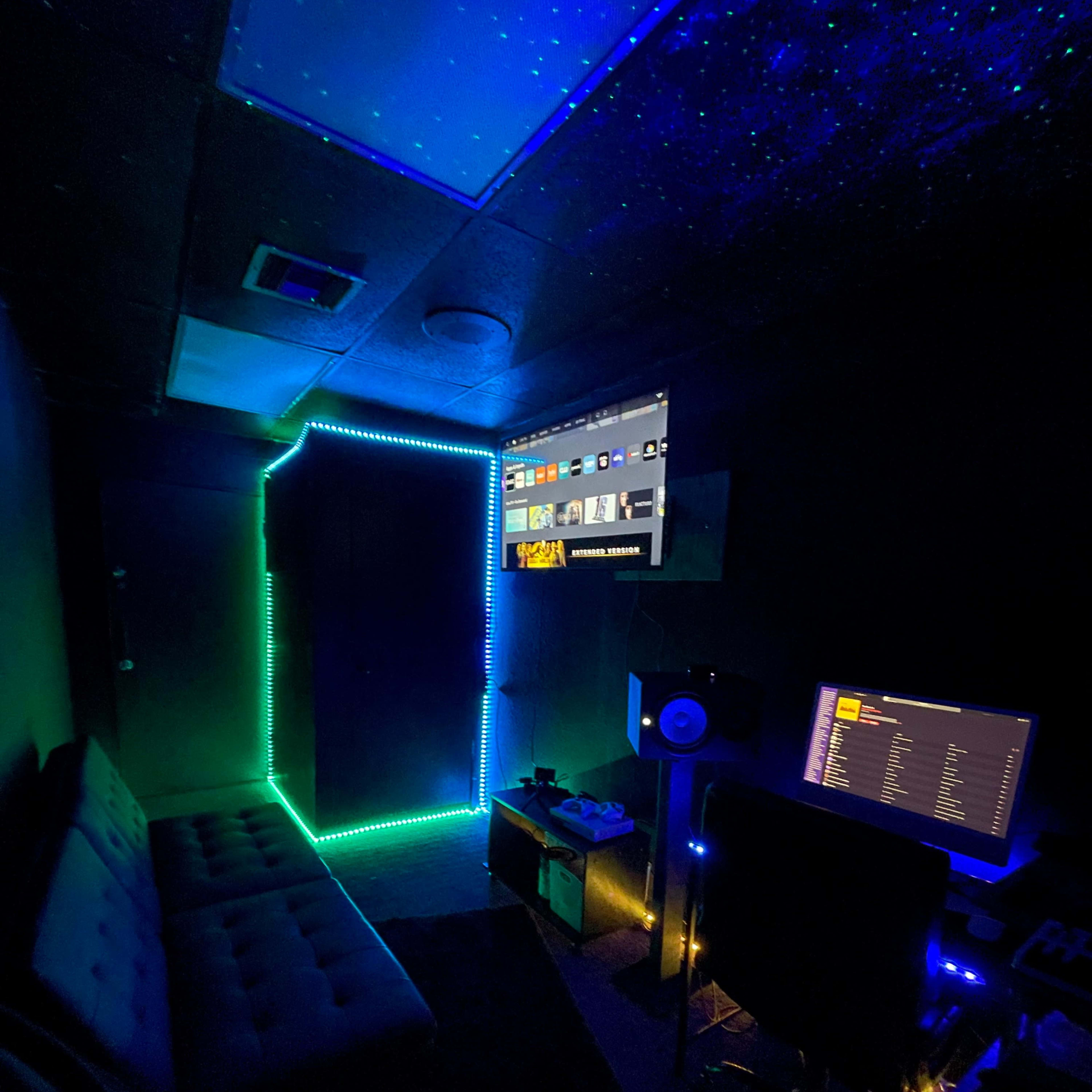 A dimly lit room with neon LED lights outlining a door and surrounding a couch, along with a computer setup and a wall-mounted screen.