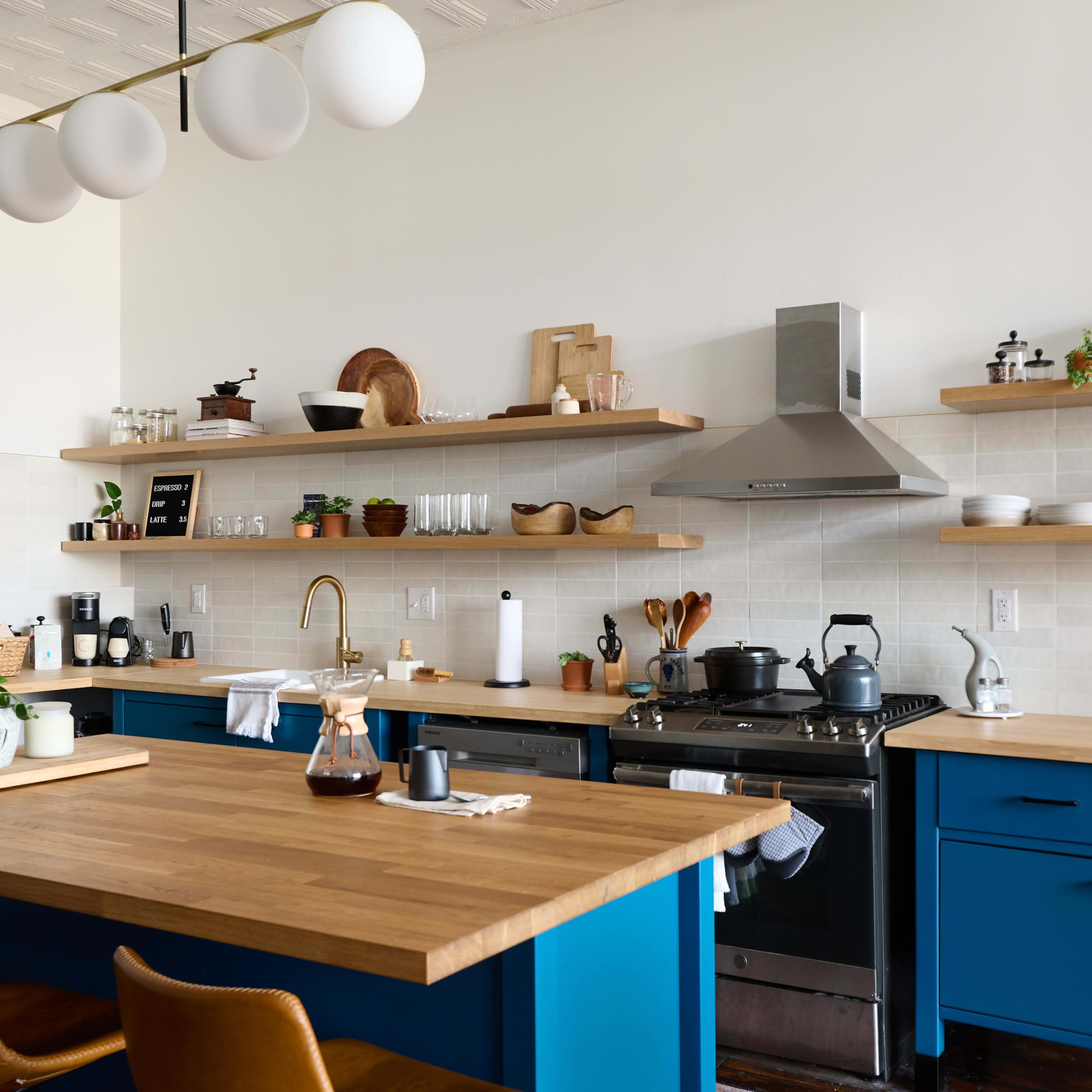 A modern kitchen features a central wooden island with blue cabinetry, open shelving displaying dishes and plants, and stainless steel appliances.