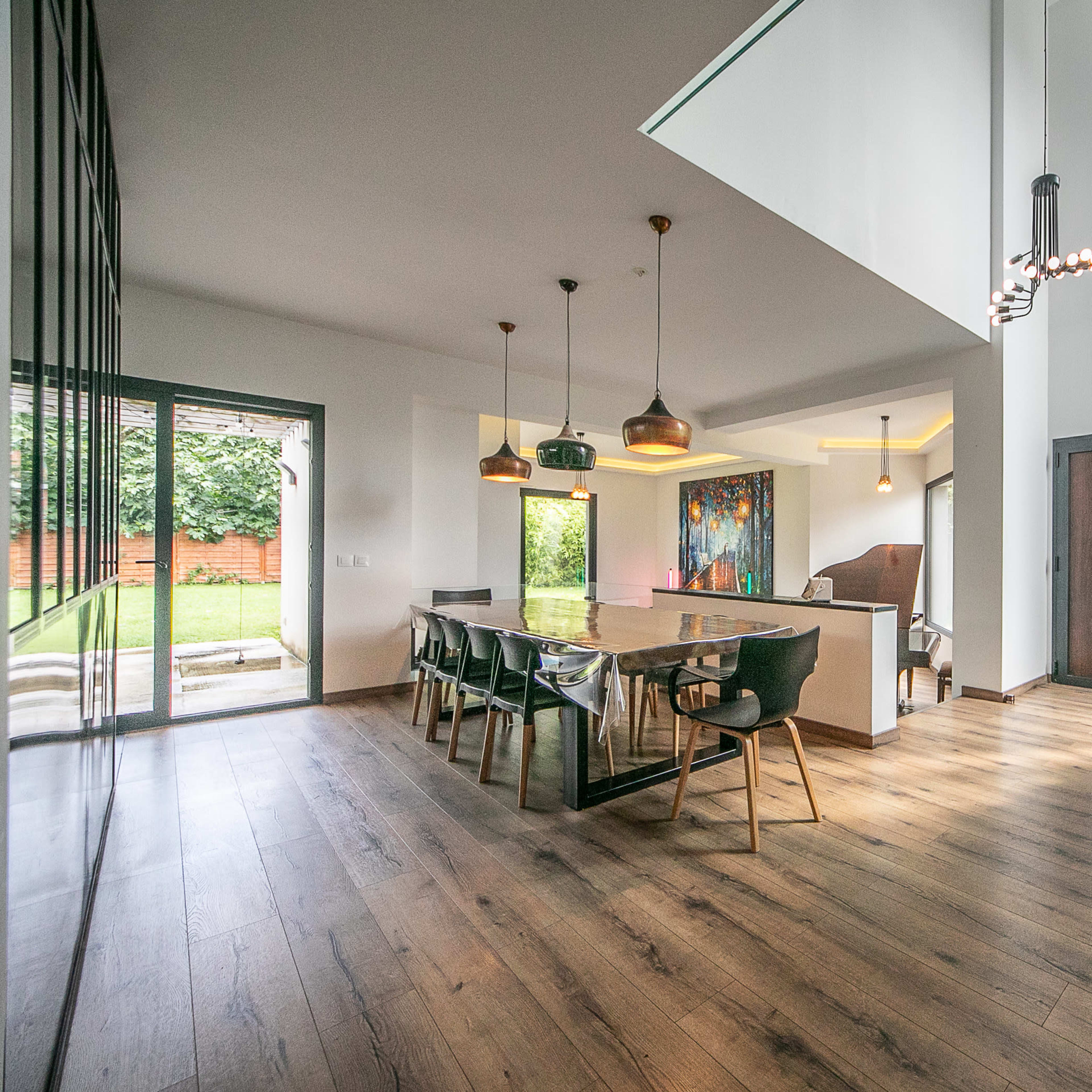 A modern living space featuring a dining area with a large table, chairs, and a kitchen, all connected to a staircase and large windows overlooking a garden.