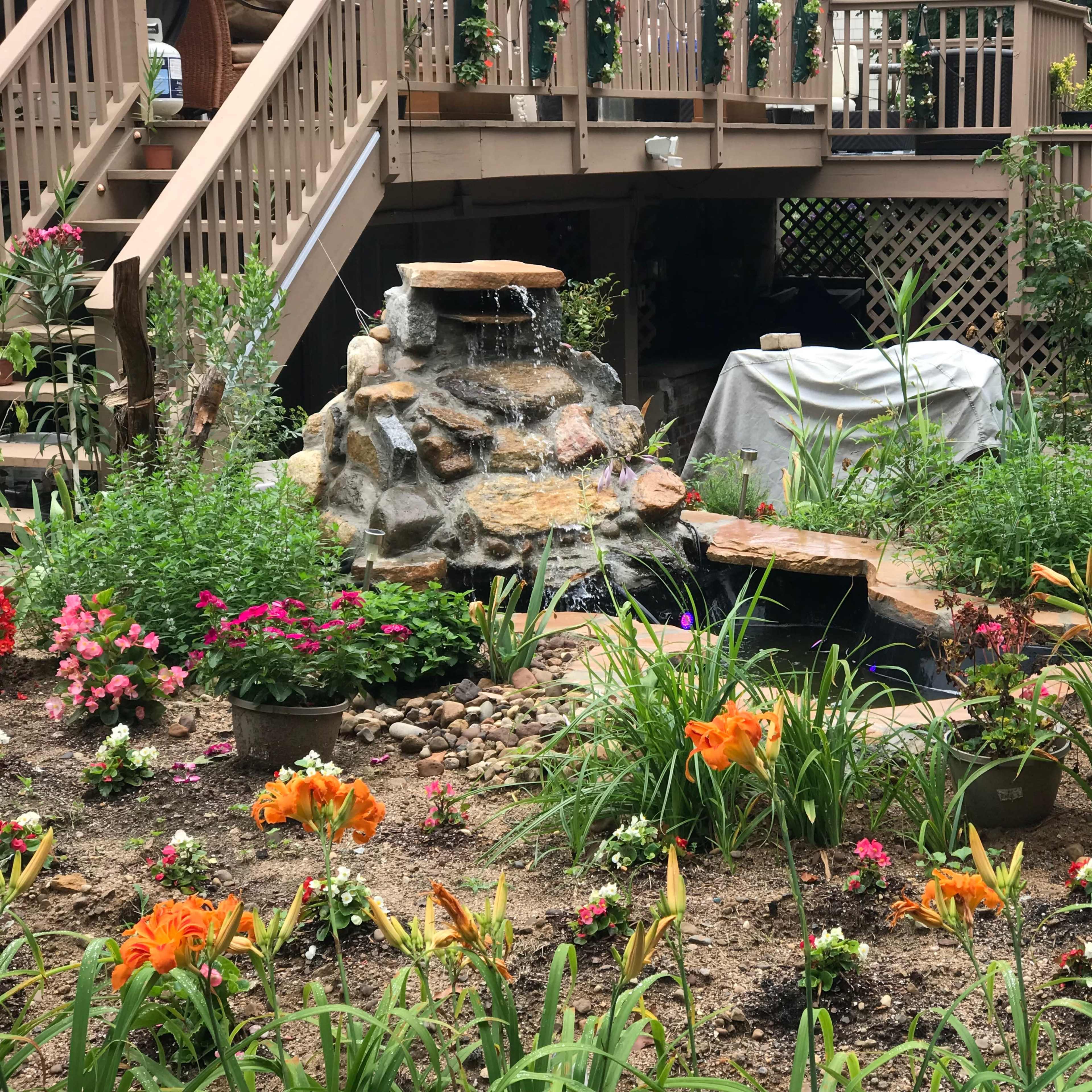 A rock garden with a waterfall is surrounded by colorful flowers and greenery beneath a wooden deck.