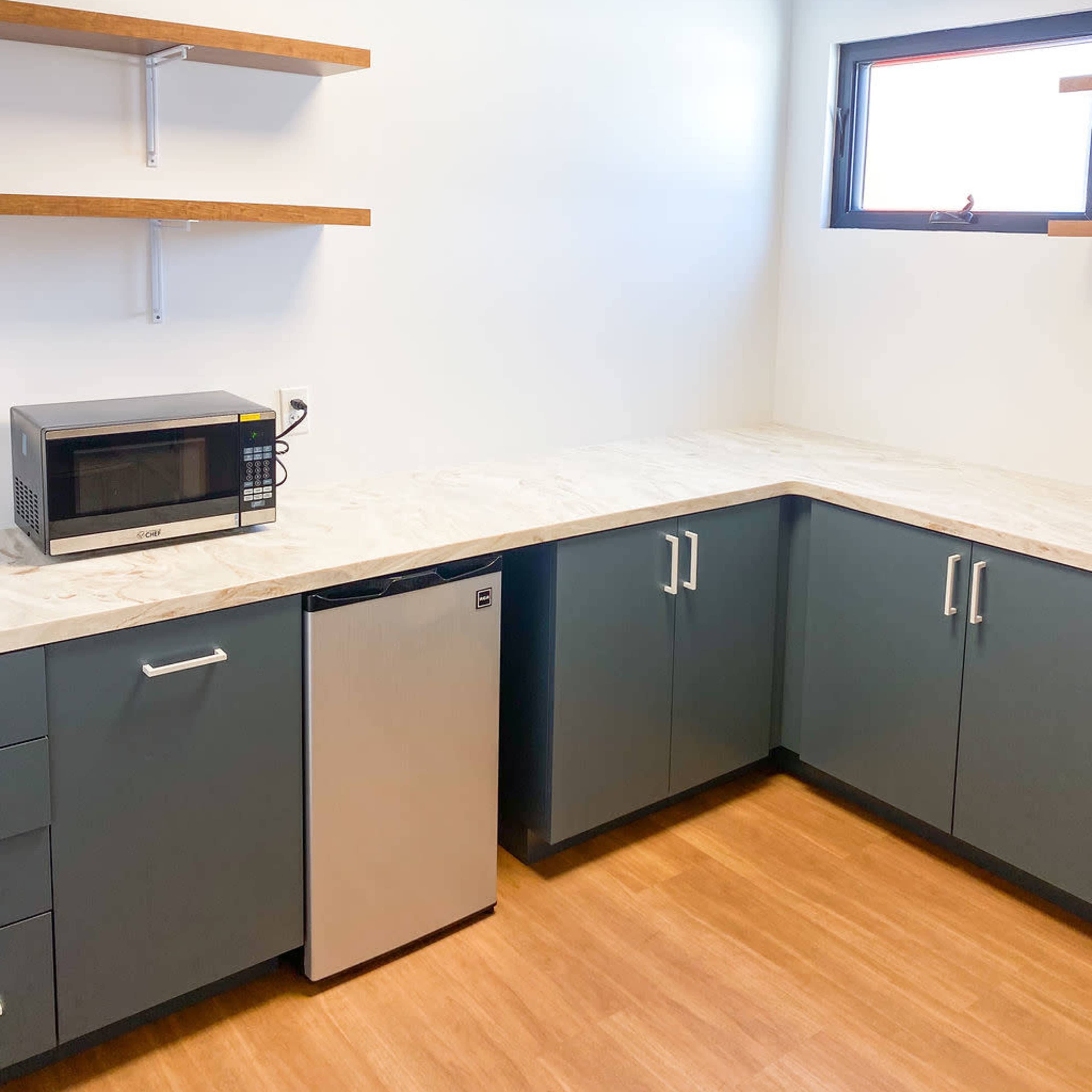 A modern kitchen area featuring a corner countertop with a microwave, a small refrigerator, and open shelves against a white wall.