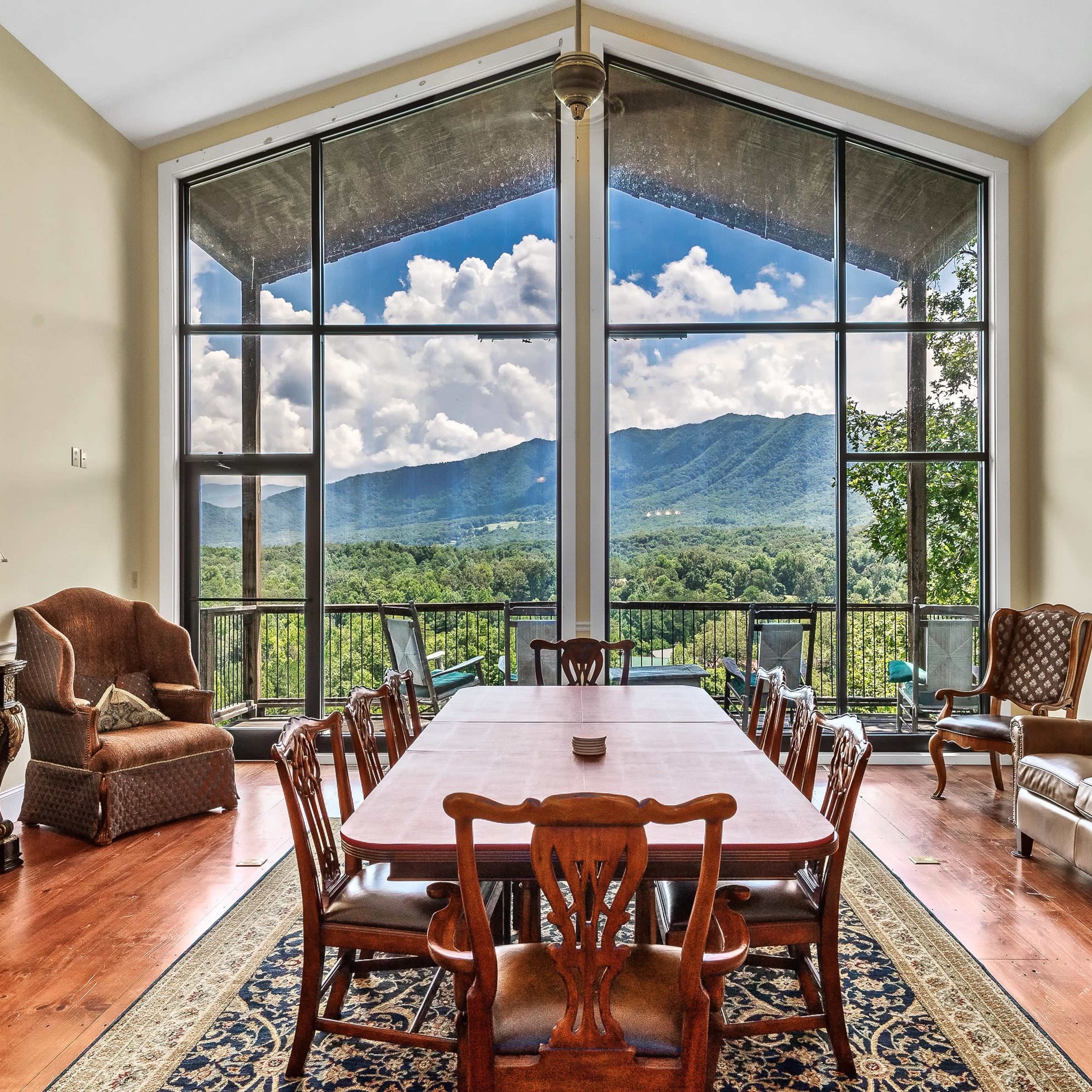 A spacious dining area with a long wooden table is surrounded by large windows that offer a view of the mountains and sky.