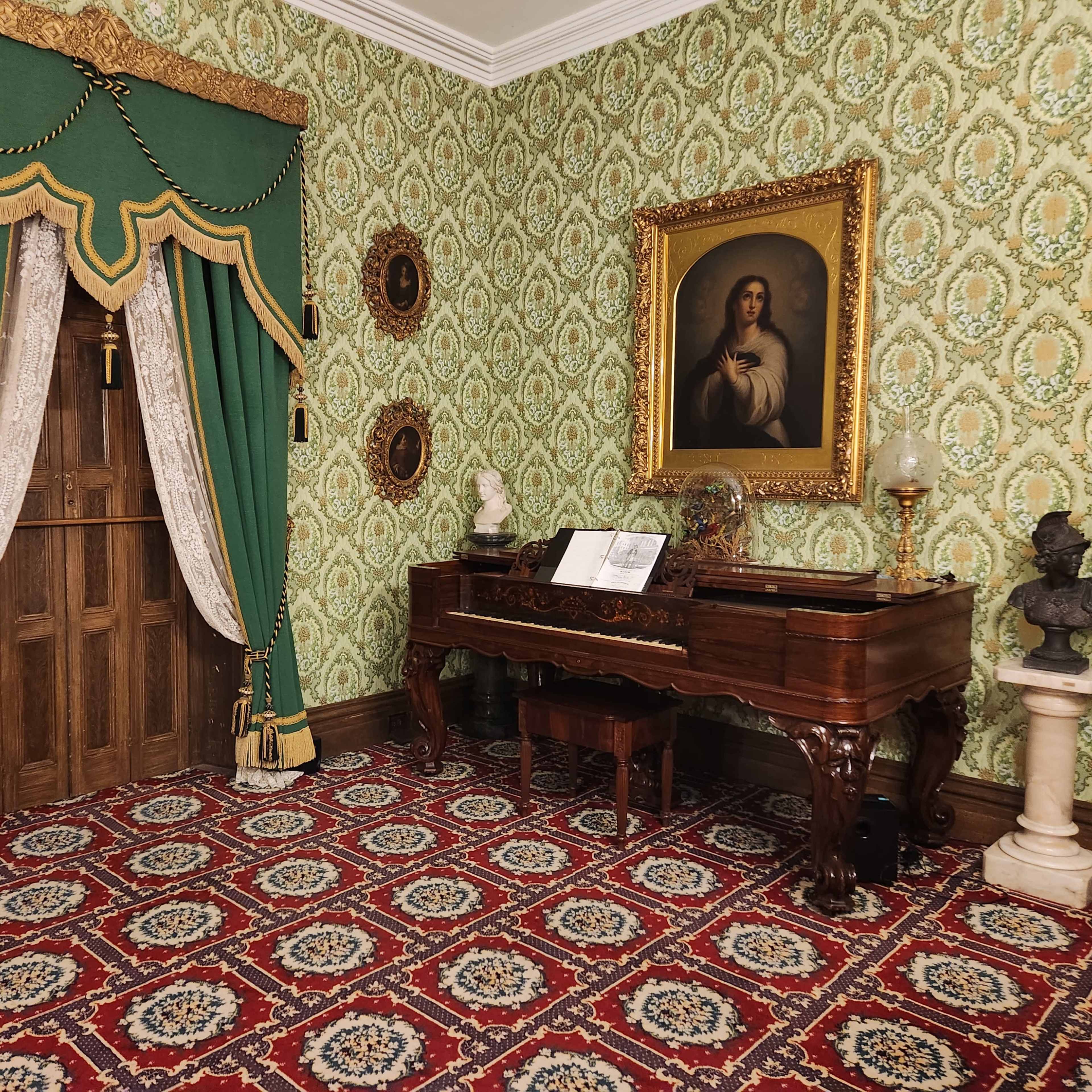 The room features green draperies framing a wooden door, a grand piano with sheet music, and a large portrait on the wall, set against ornate wallpaper and a patterned carpet.