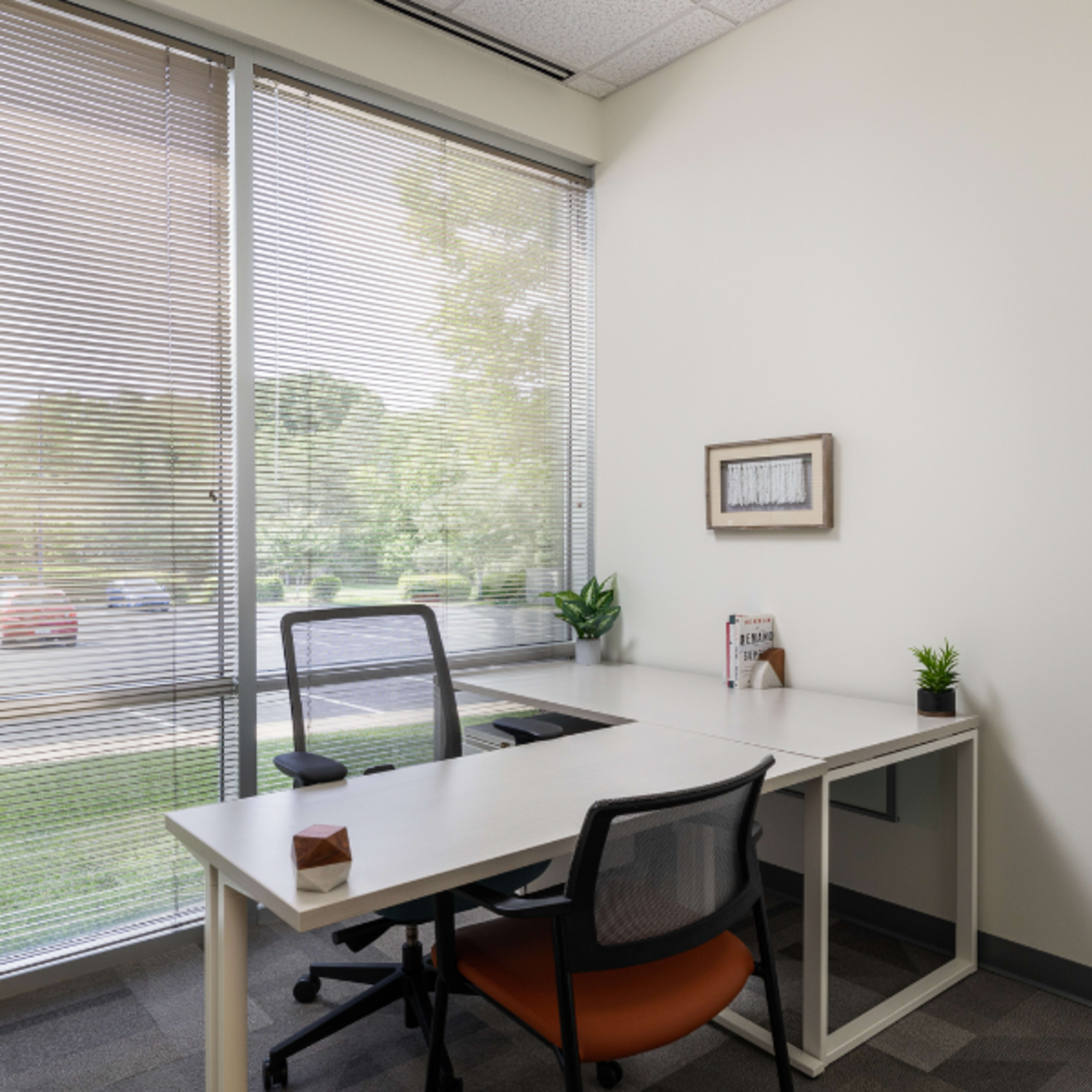 A modern office space with a desk, two chairs, and a large window featuring horizontal blinds.