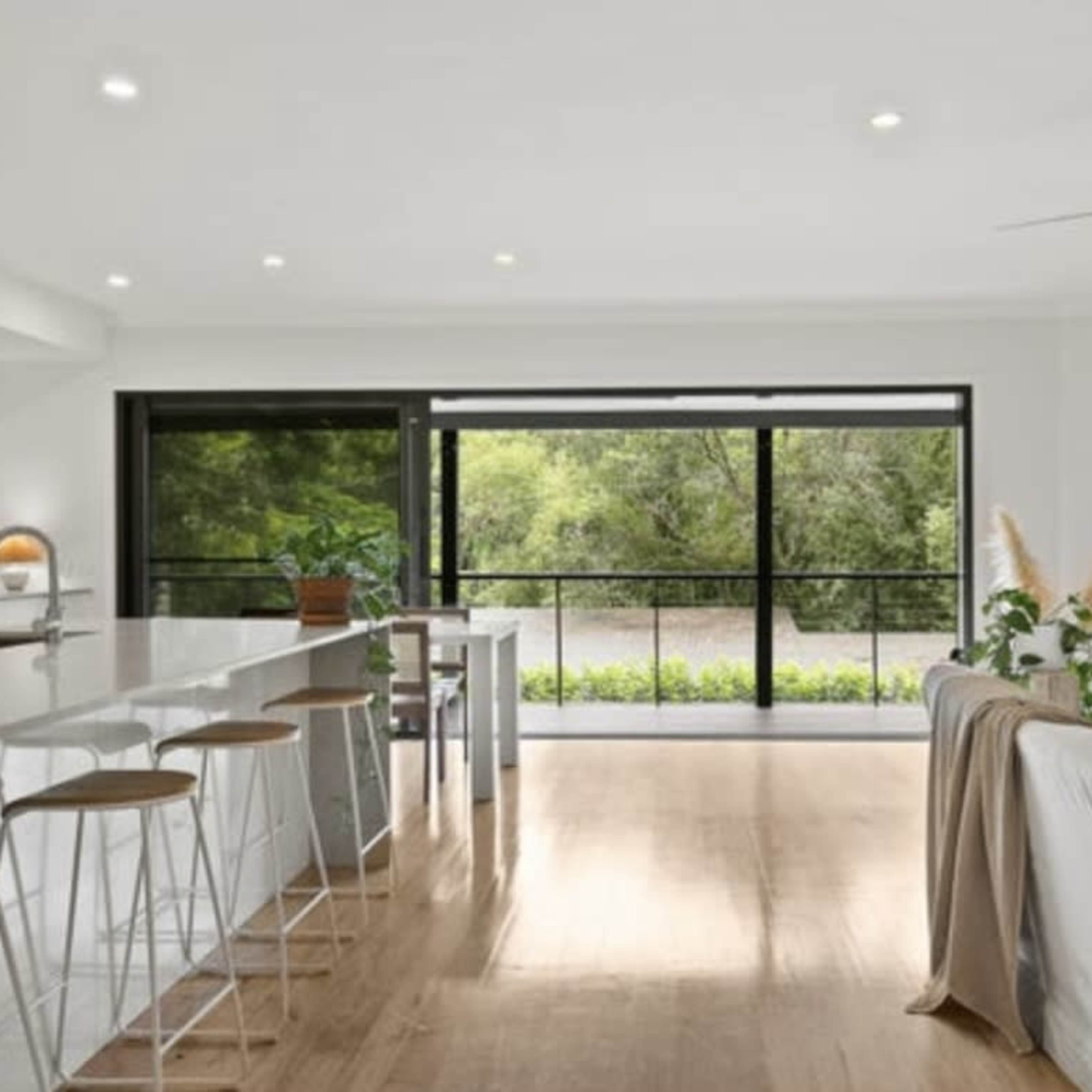 A modern living space features a spacious kitchen with a long island bar, large windows overlooking greenery, and a comfortable seating area.