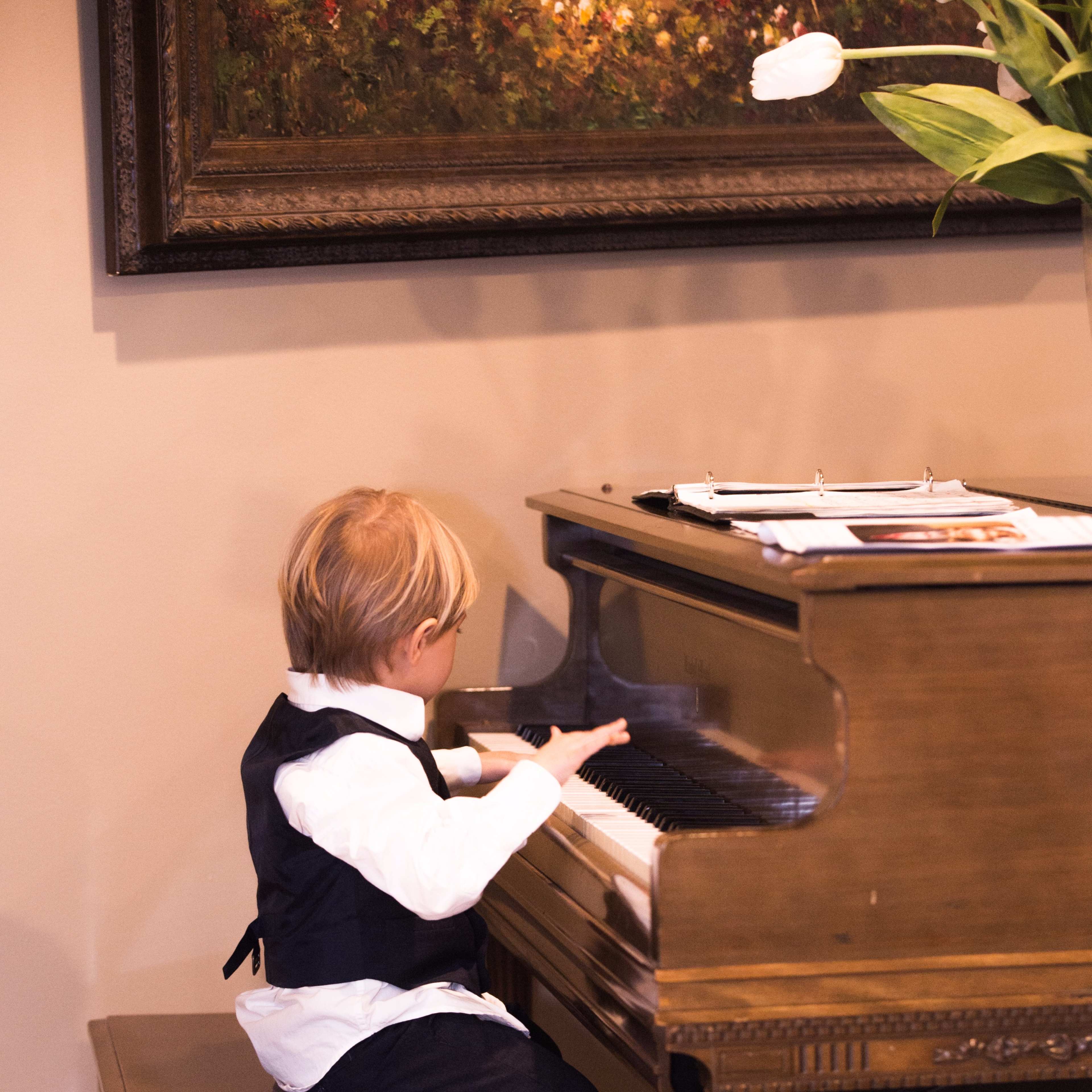 A young boy in formal attire sits on a wooden stool, playing a piano in a well-lit room.