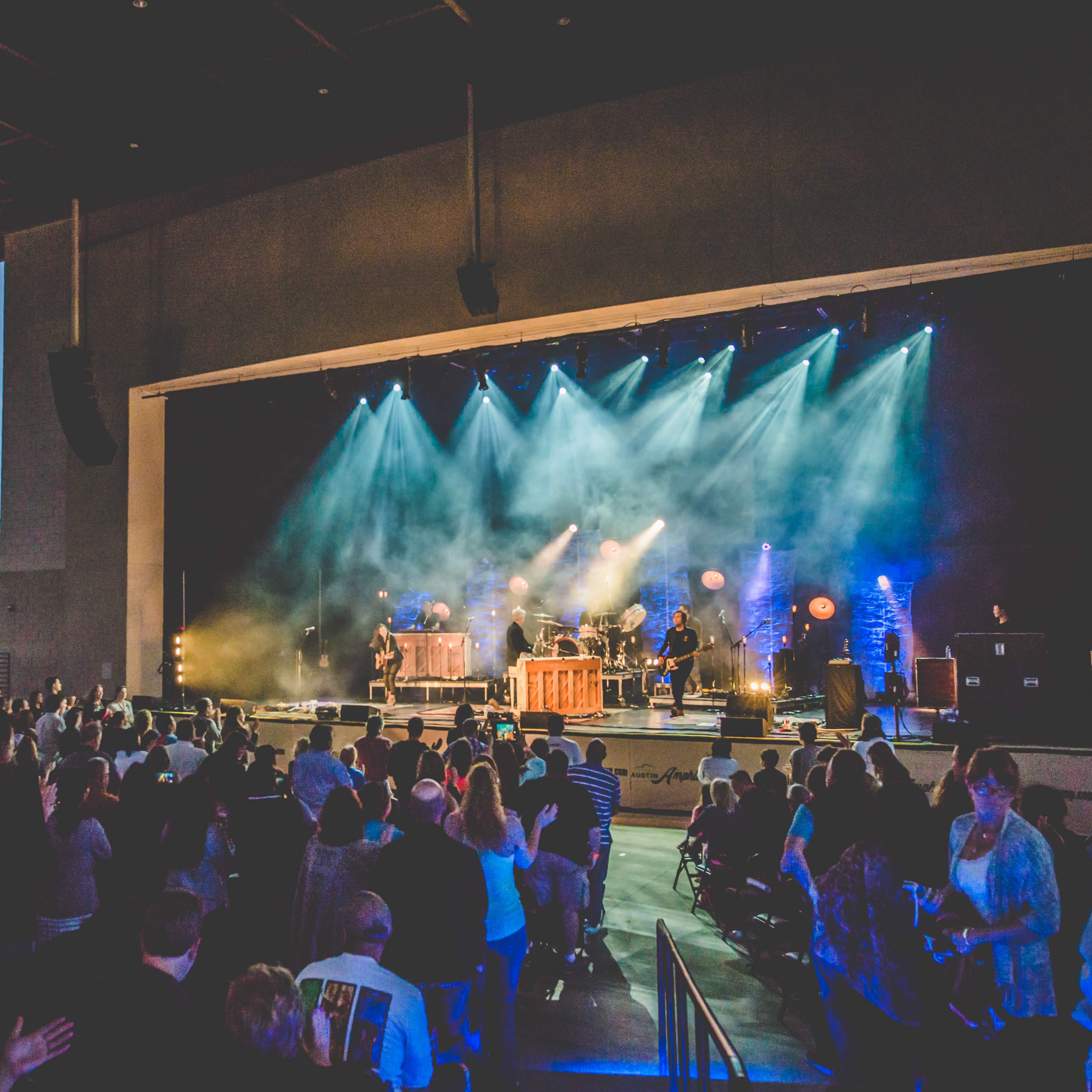 A live music concert is taking place on an outdoor stage with a crowd watching and colorful lights illuminating the performers.