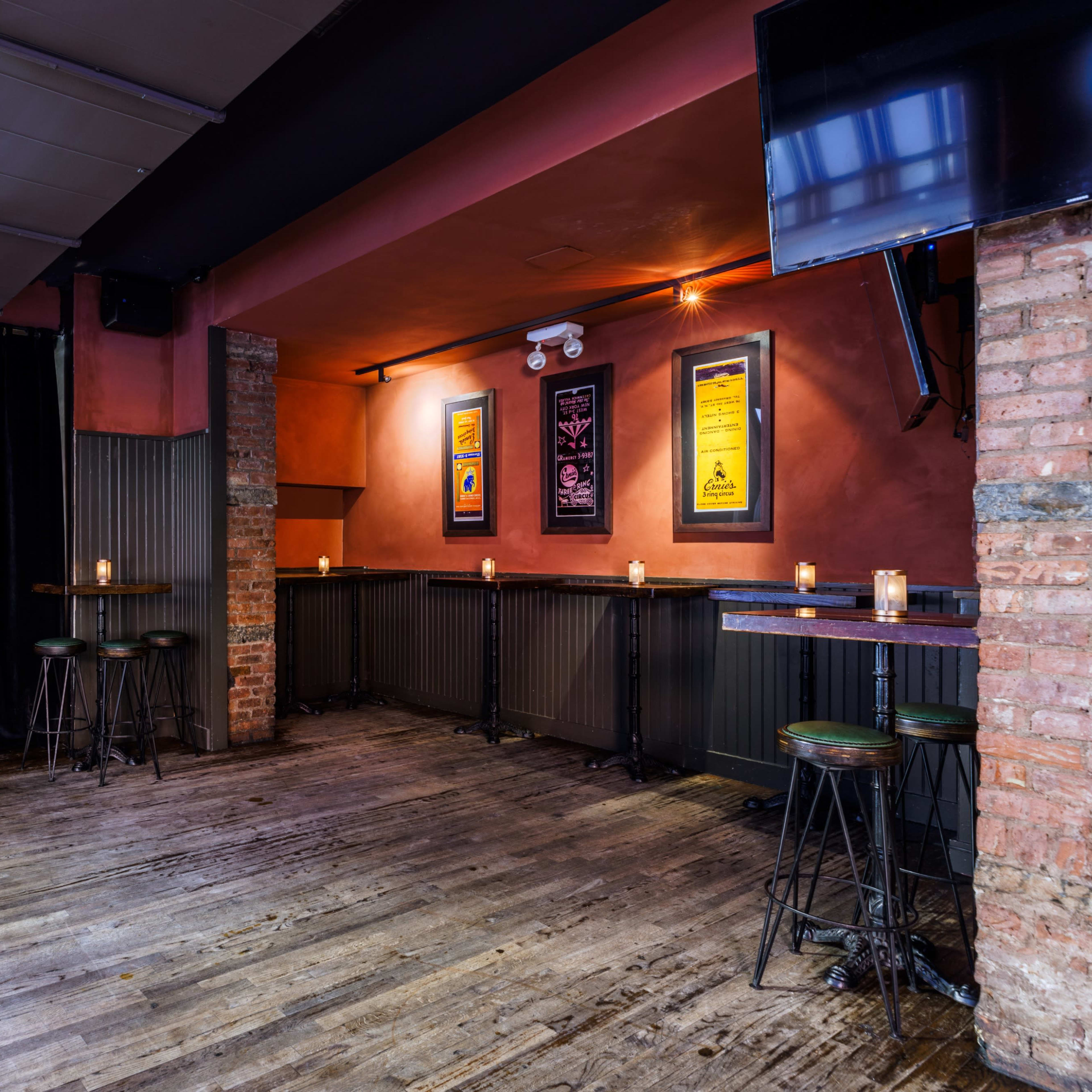 A dimly lit bar area with wooden tables, metal stools, and candlelight on the tables, featuring brick and painted walls decorated with posters.