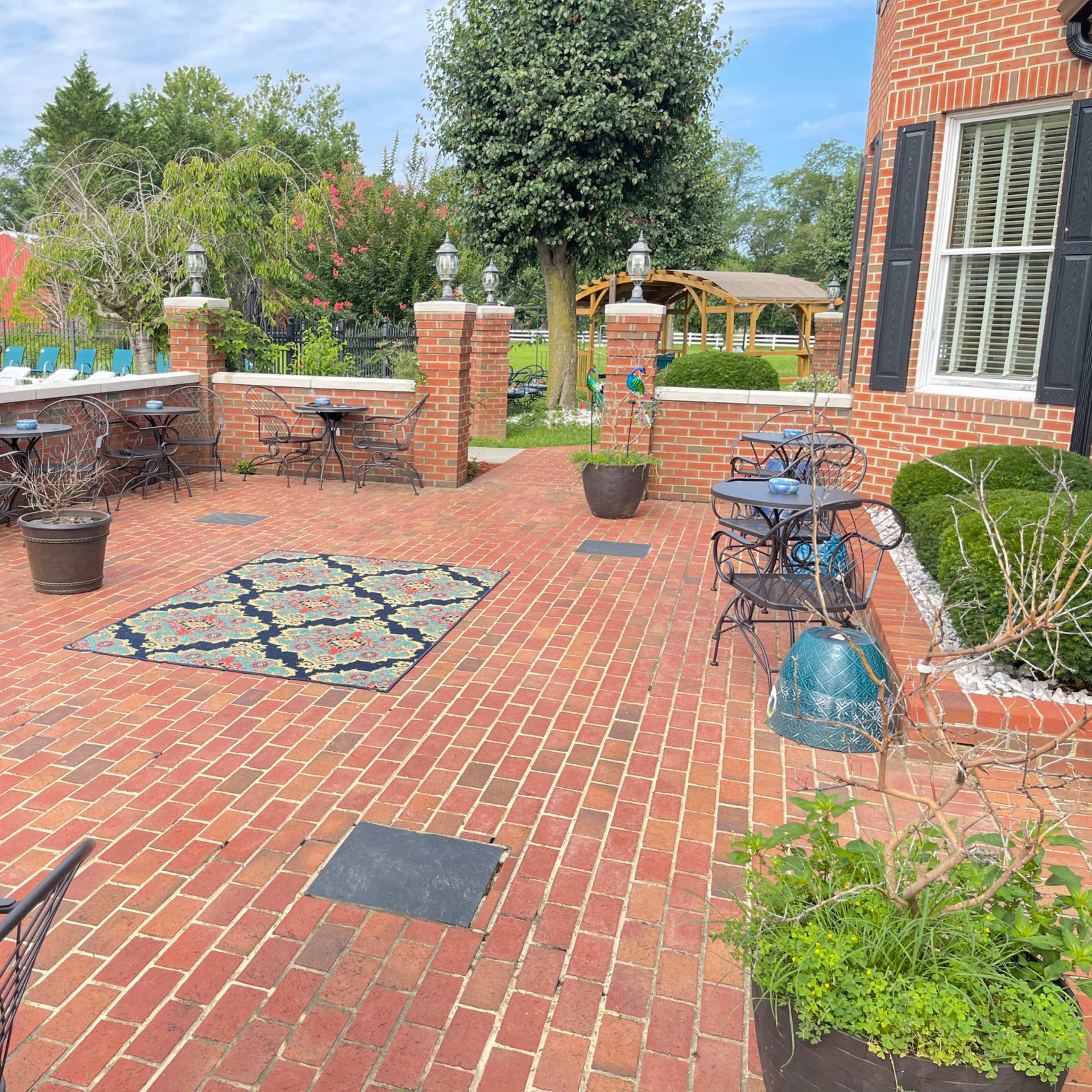 A brick patio with several black metal tables and chairs, a decorative rug, and surrounding greenery.