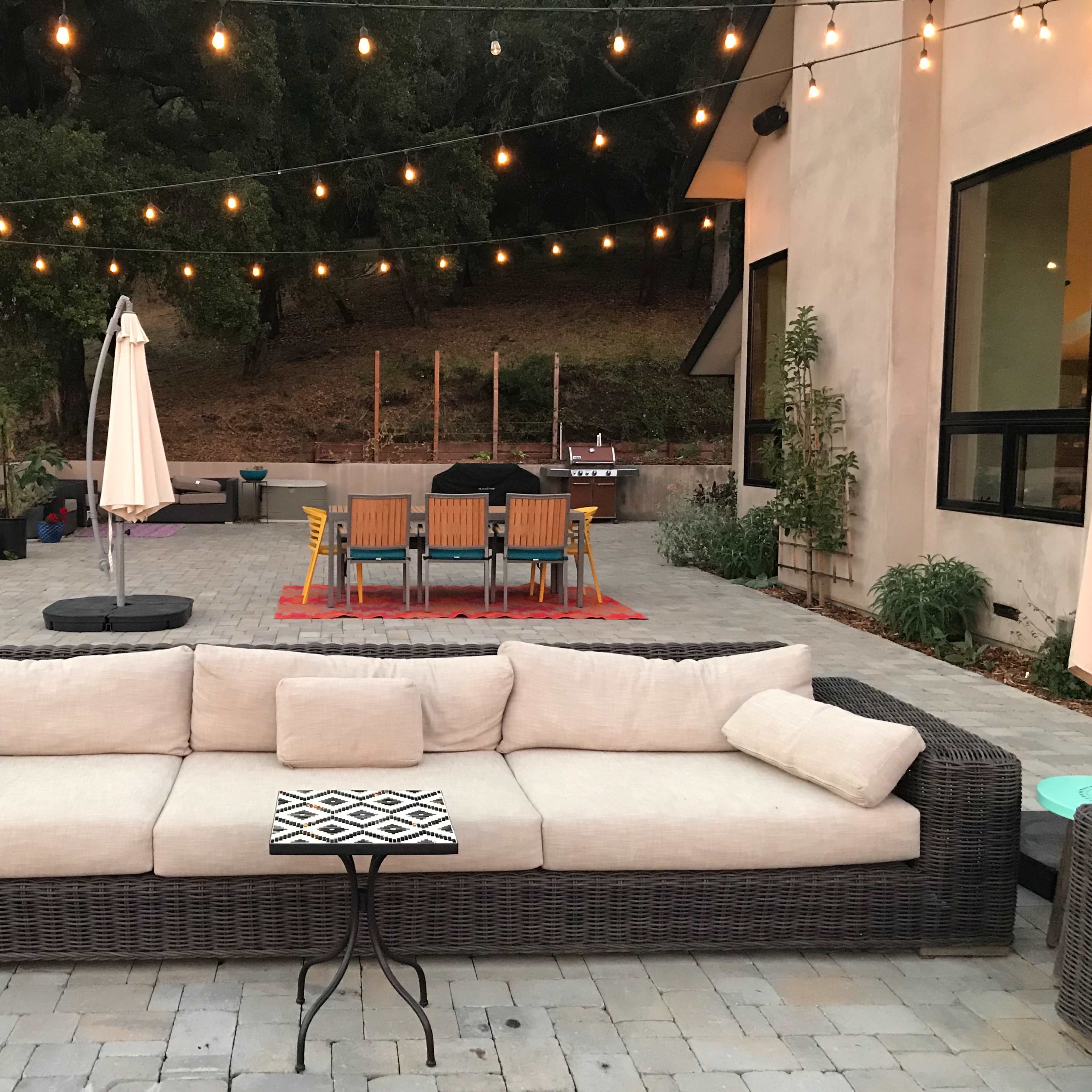 A patio area featuring a large wicker sofa with a small table, a dining set in the background, and string lights overhead.