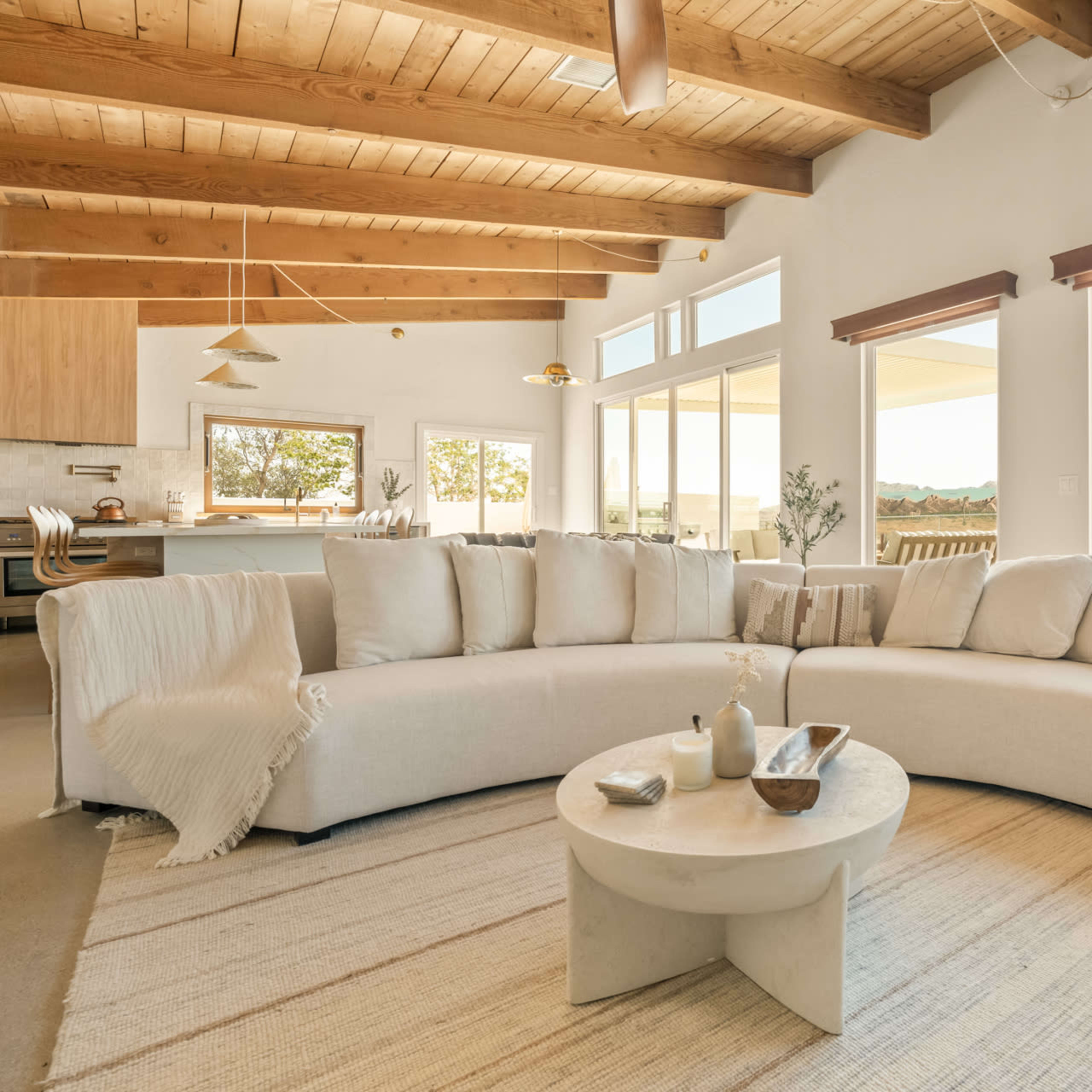 A spacious living area features a curved beige couch, a round coffee table, and large windows providing views of the outdoors.