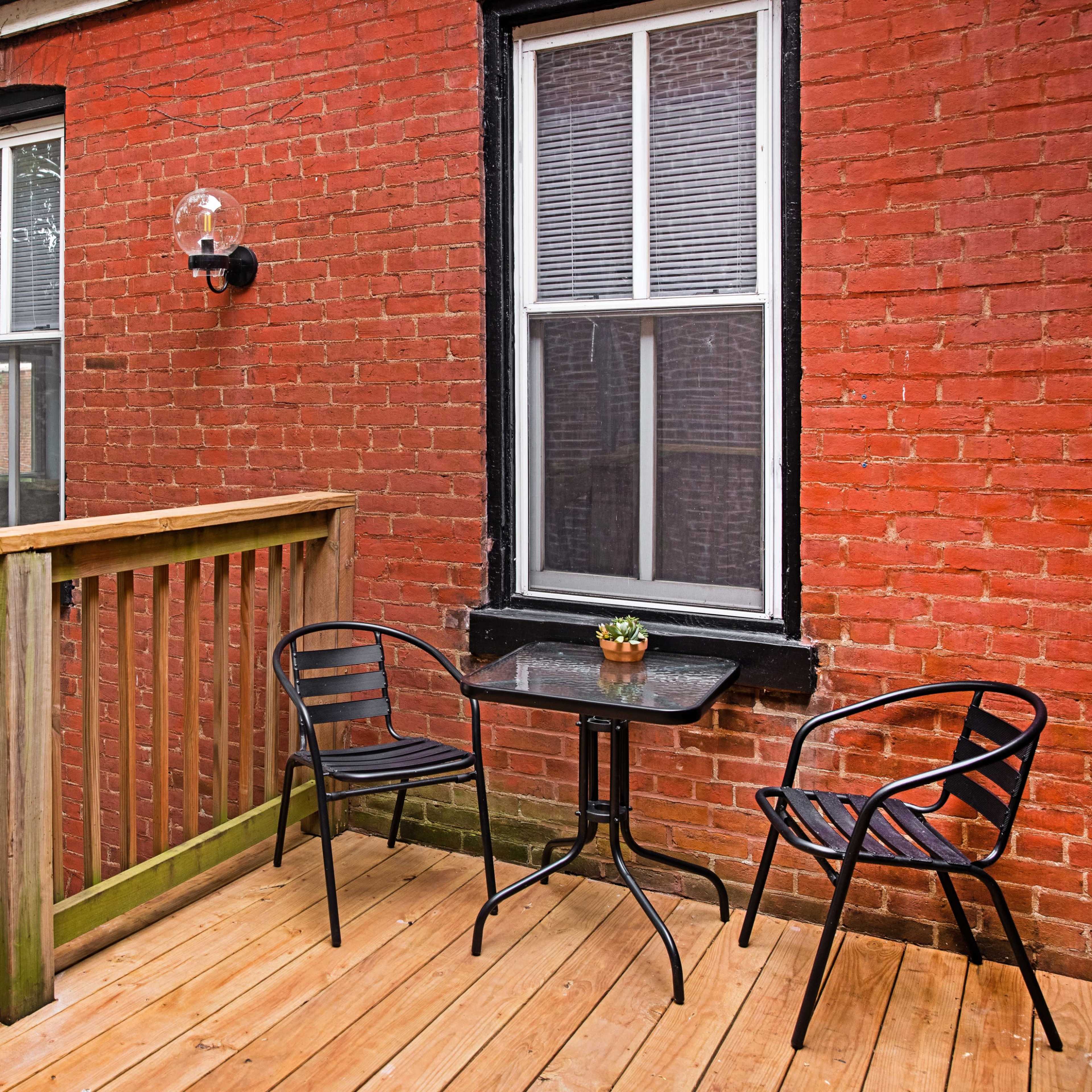 A small patio features a wooden deck, two black metal chairs, and a table with a potted plant against a brick wall.