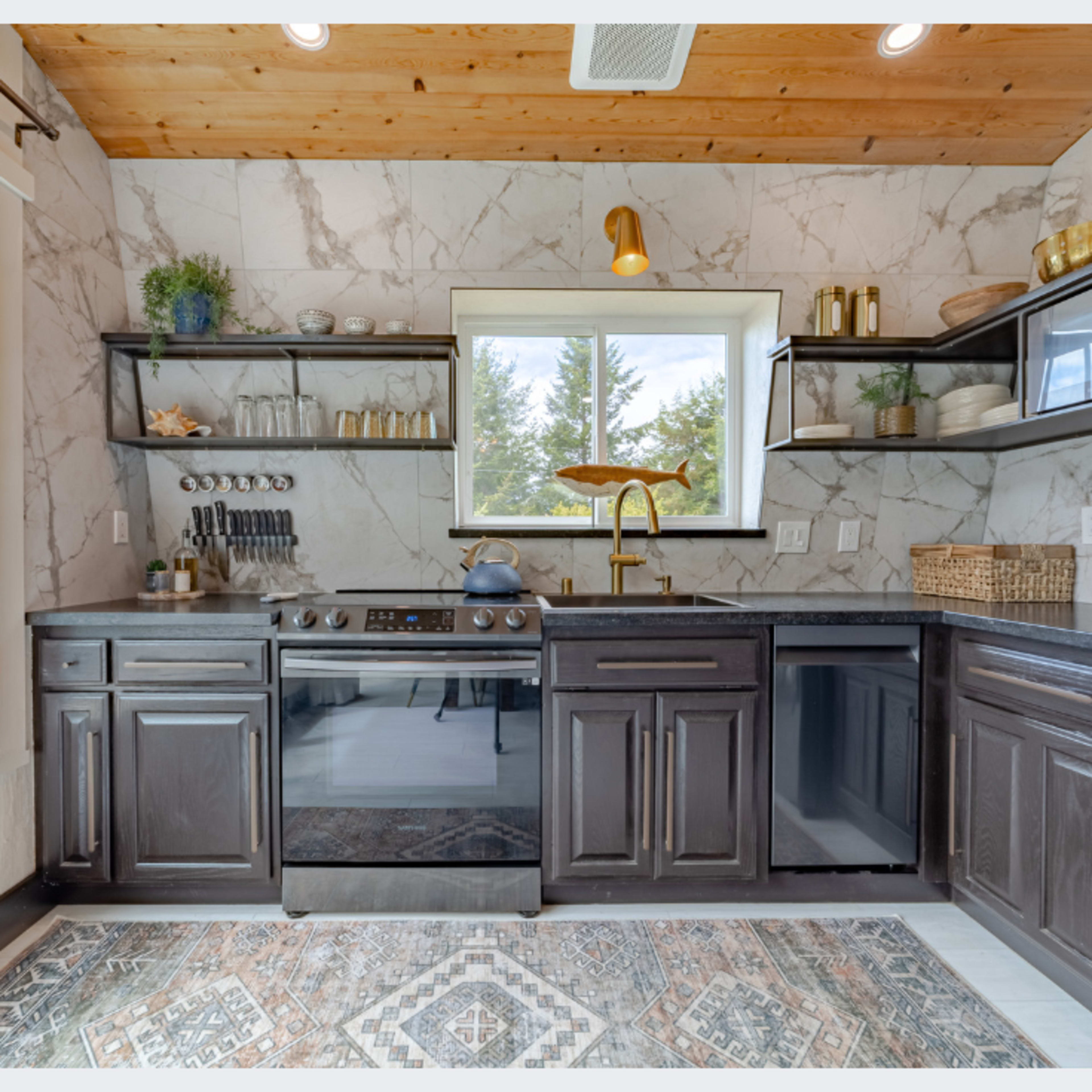 A modern kitchen with dark cabinetry, a stainless steel stove, and open shelving, featuring a window that offers a view of the outdoors.