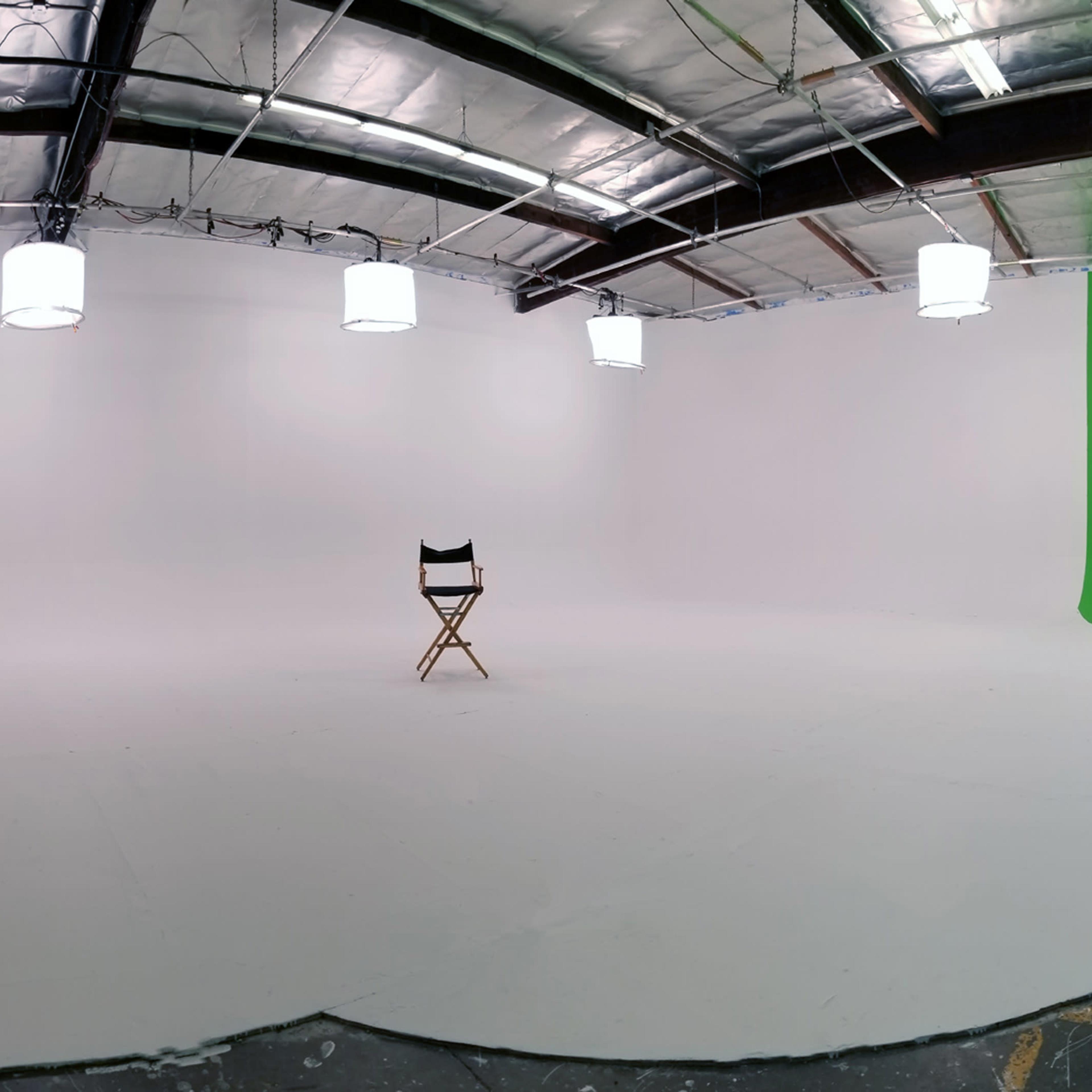 A large, empty studio space with a wooden director's chair centered on a white floor, surrounded by hanging lights and green and black backdrops.