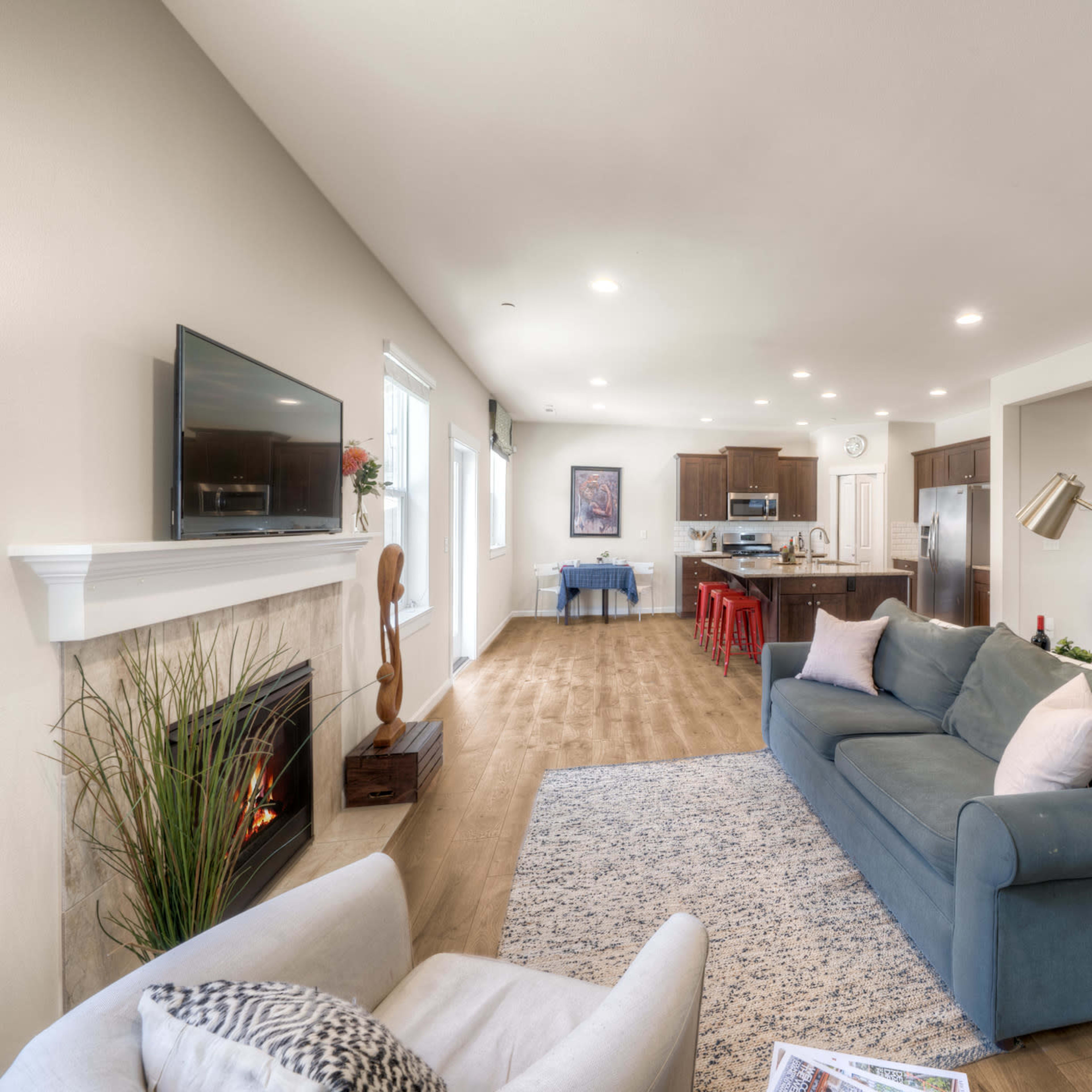 A modern living room with a blue couch, a fireplace, and an open layout connecting to a kitchen with a dining area in the background.