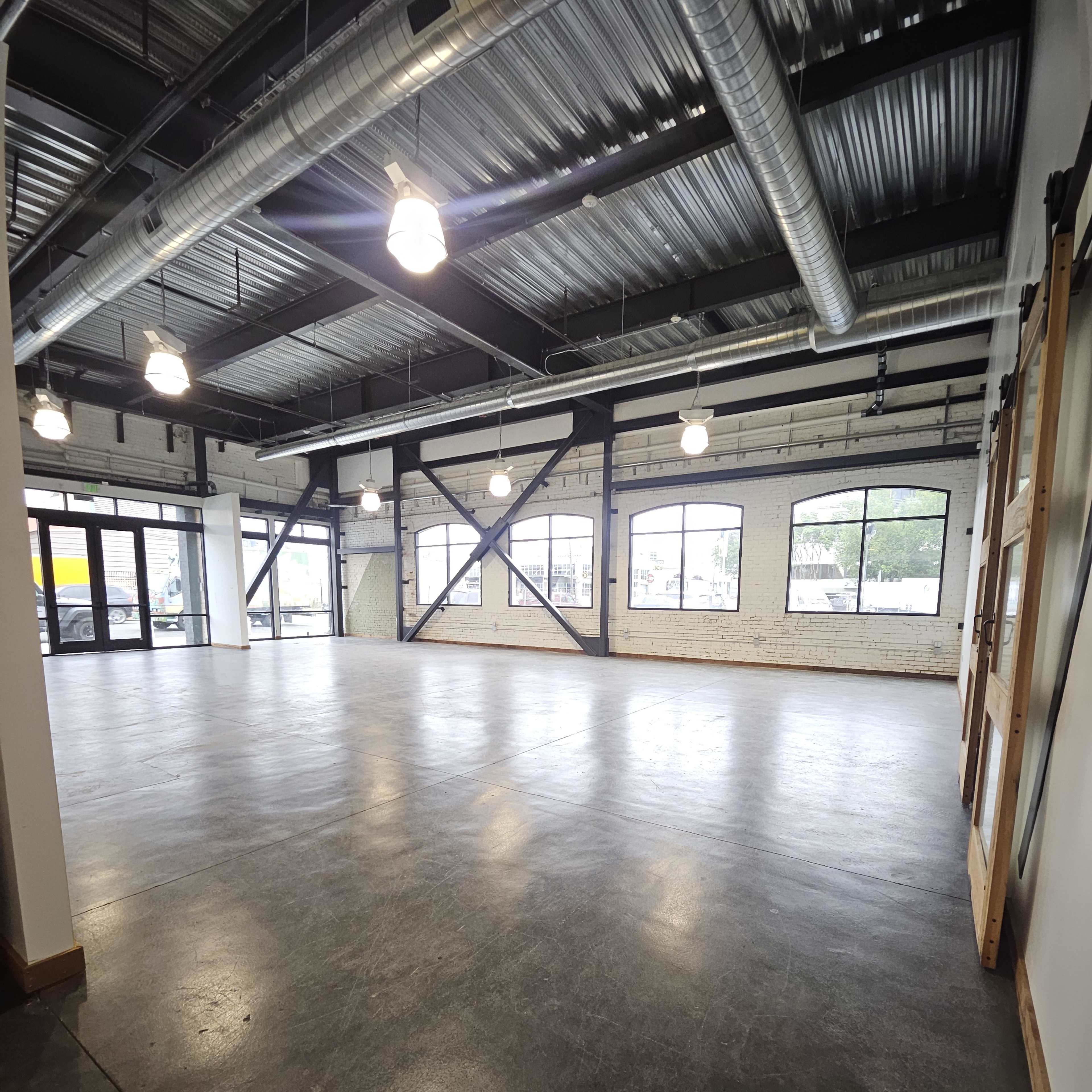 An empty industrial-style interior space with concrete flooring, exposed beams, and large windows.