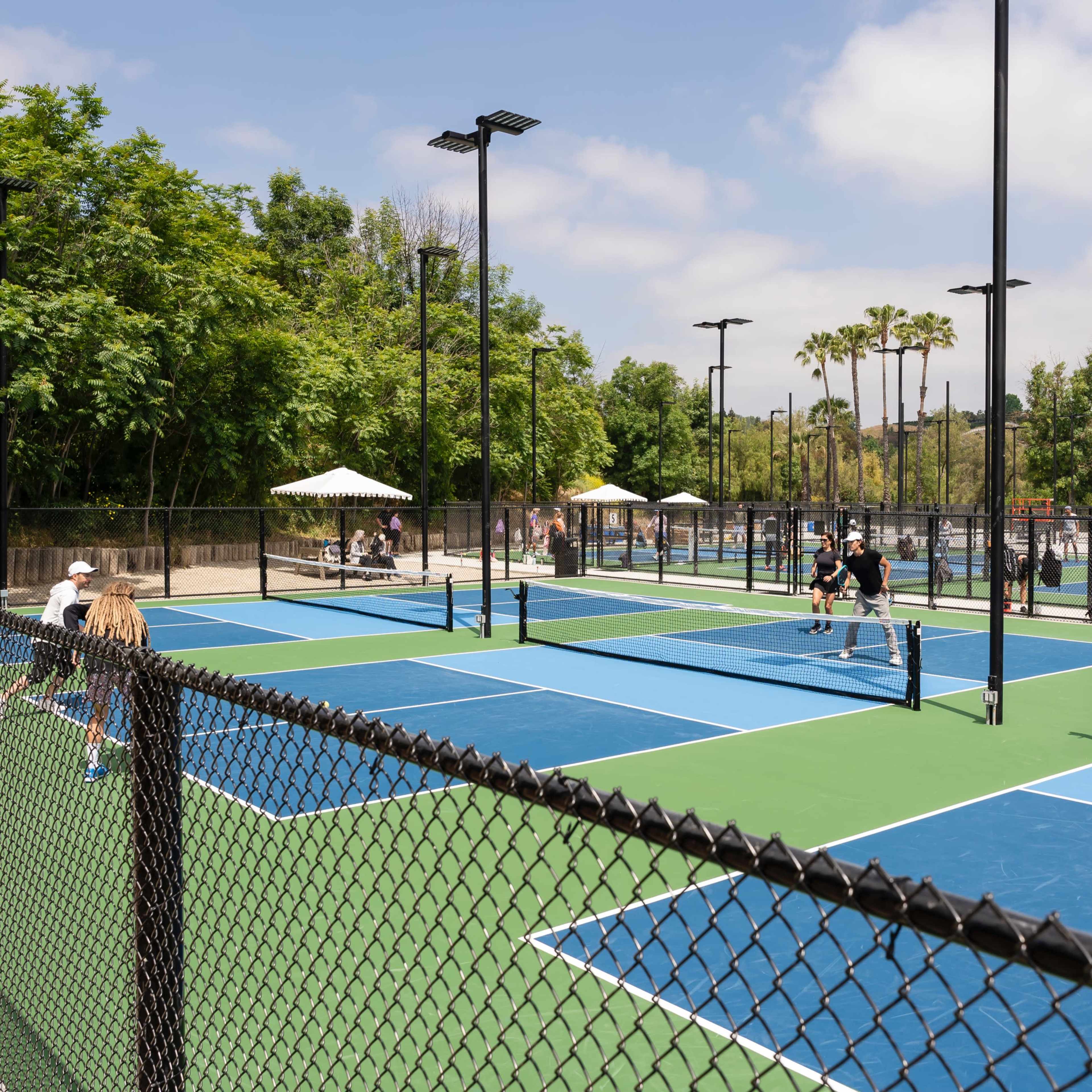 A vibrant outdoor pickleball complex with multiple courts, players engaged in games, and spectators sitting under umbrellas in a landscaped area.