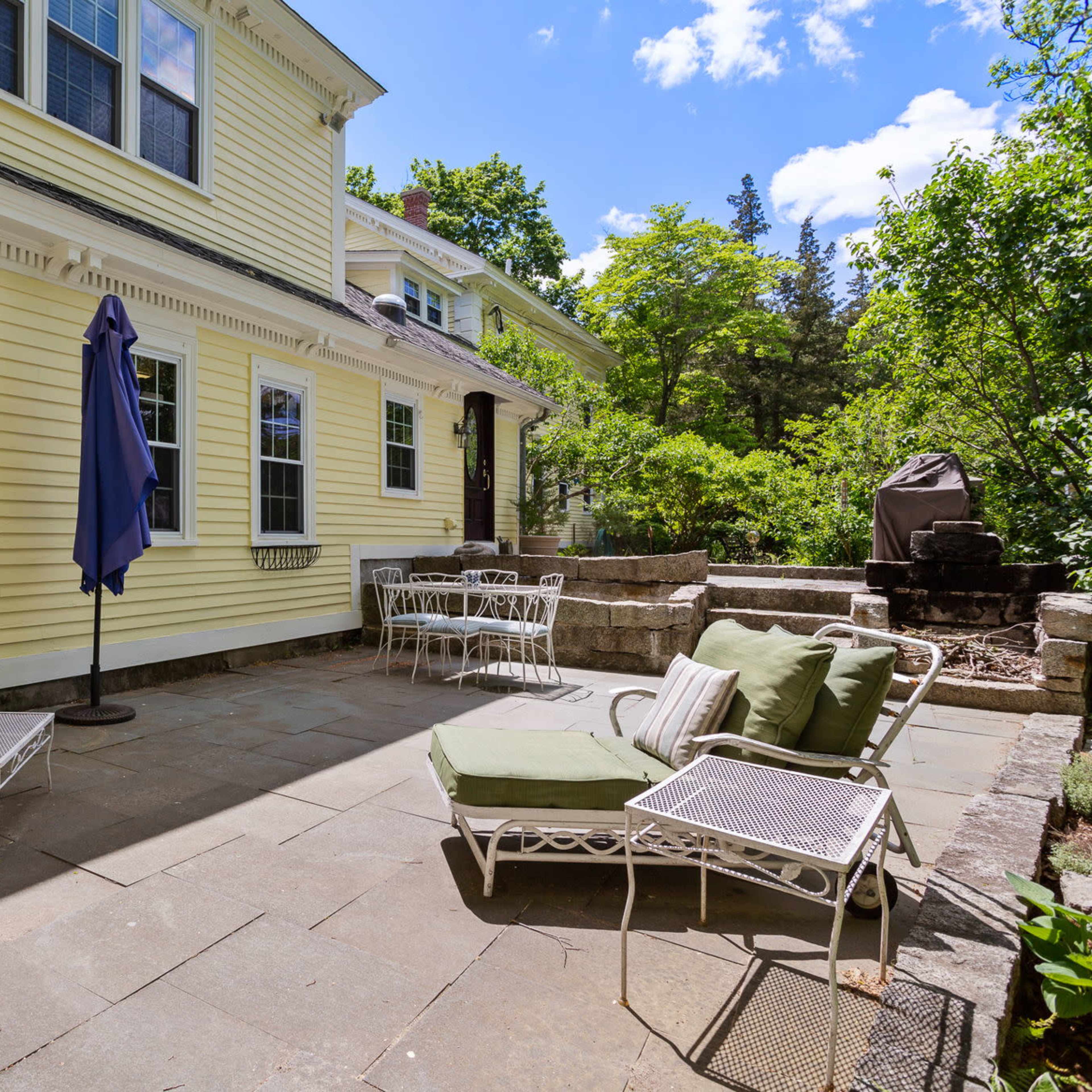 A patio area with a variety of outdoor furniture, including lounge chairs and a small table, adjacent to a yellow house and surrounded by greenery.