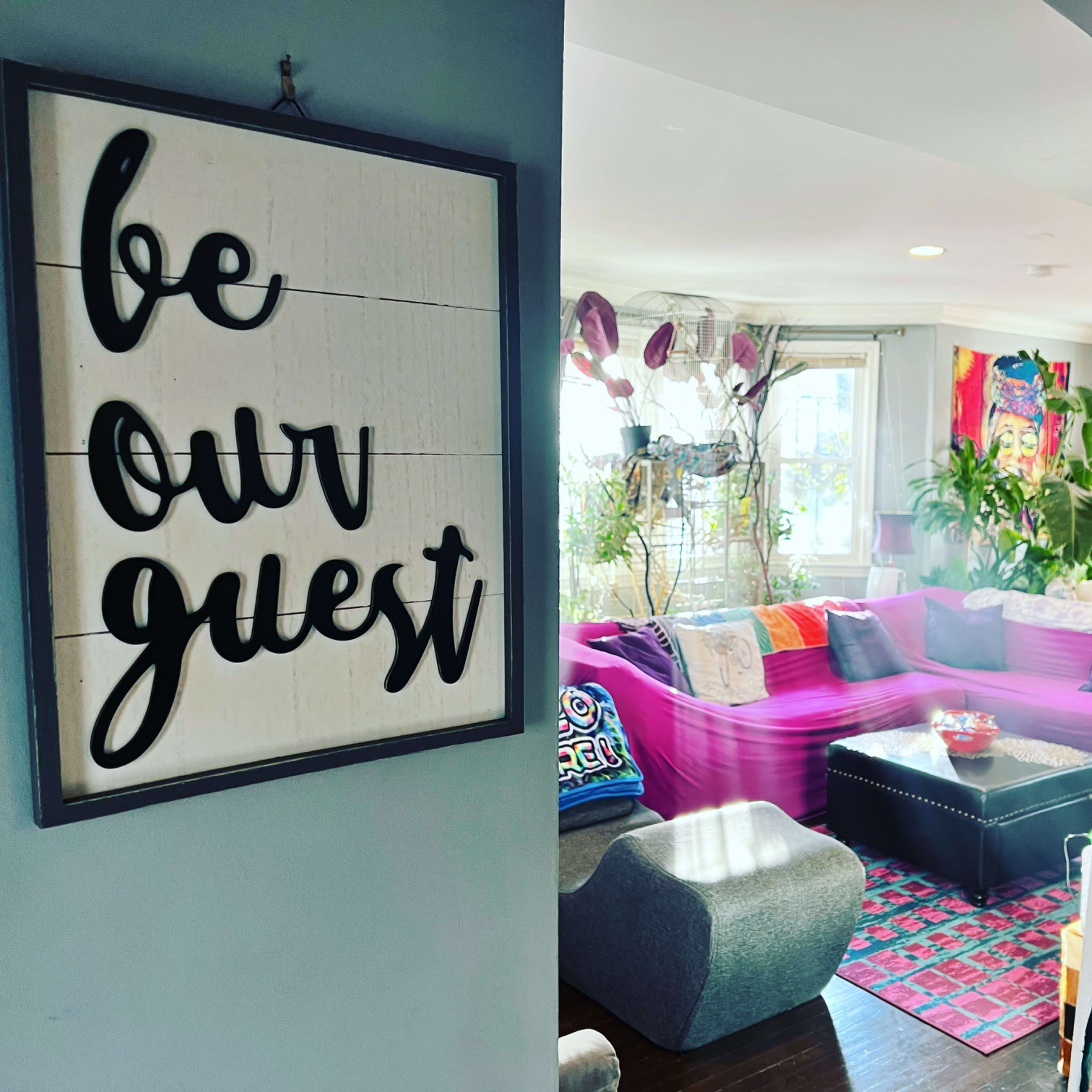 A welcoming living room with vibrant decor, plants, and a sign that reads "be our guest" on the wall.