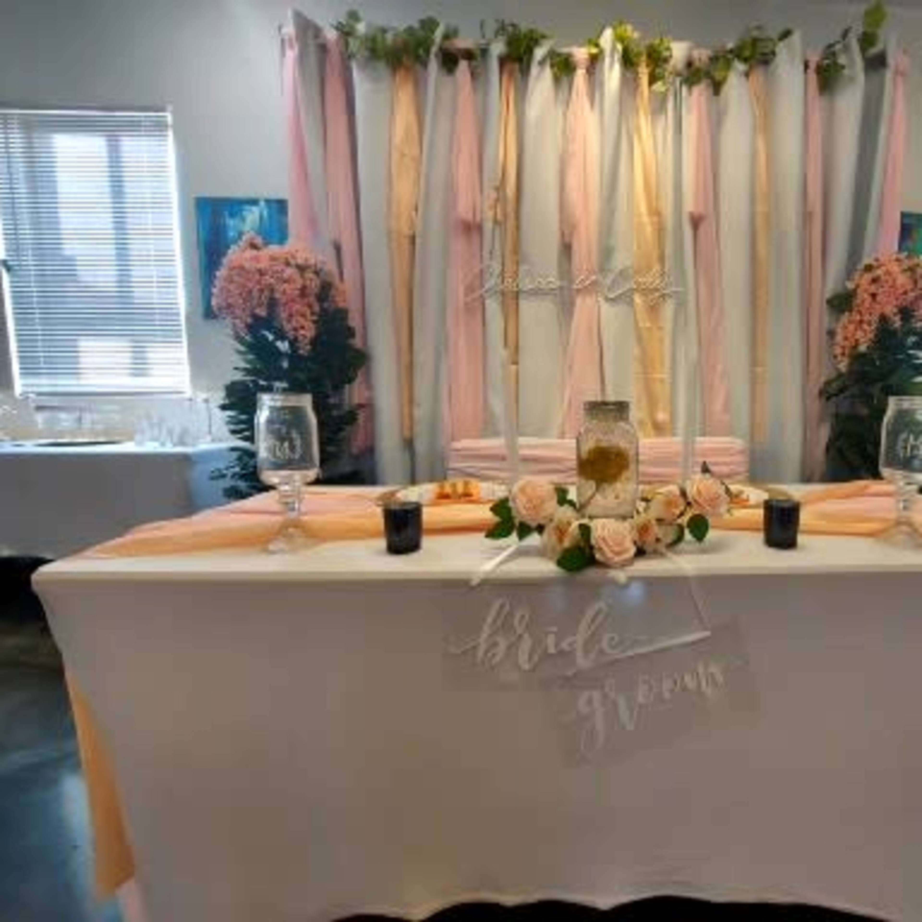 A decorated wedding reception area with a table set for the bride and groom, featuring floral arrangements and pastel drapes as a backdrop.