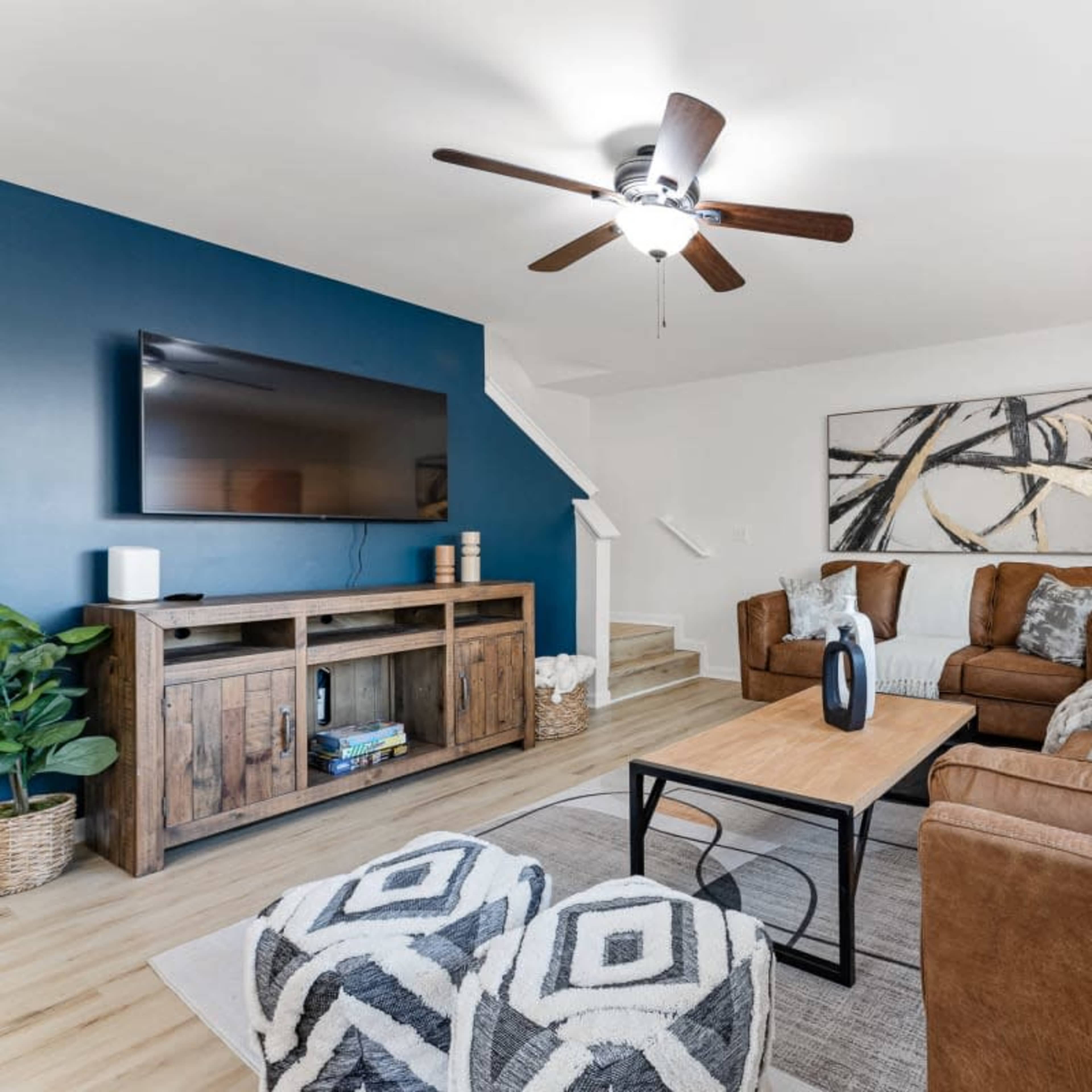 A living room features a brown suede sofa set, a wooden TV stand, and a coffee table, all arranged around a light-colored rug with patterned ottomans.