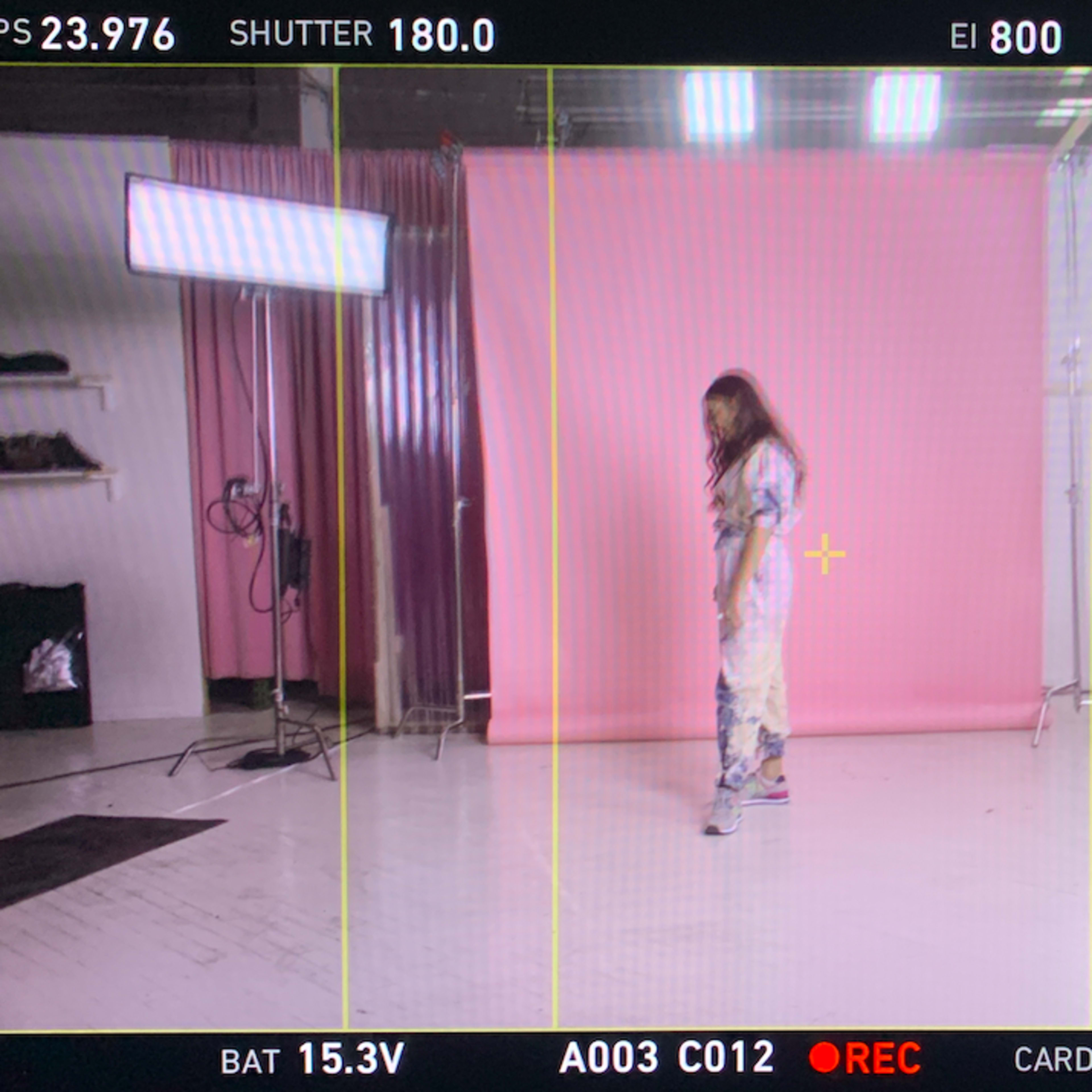 A model stands in a photography studio with a pink backdrop, bright lights, and a computer monitor displaying camera settings.