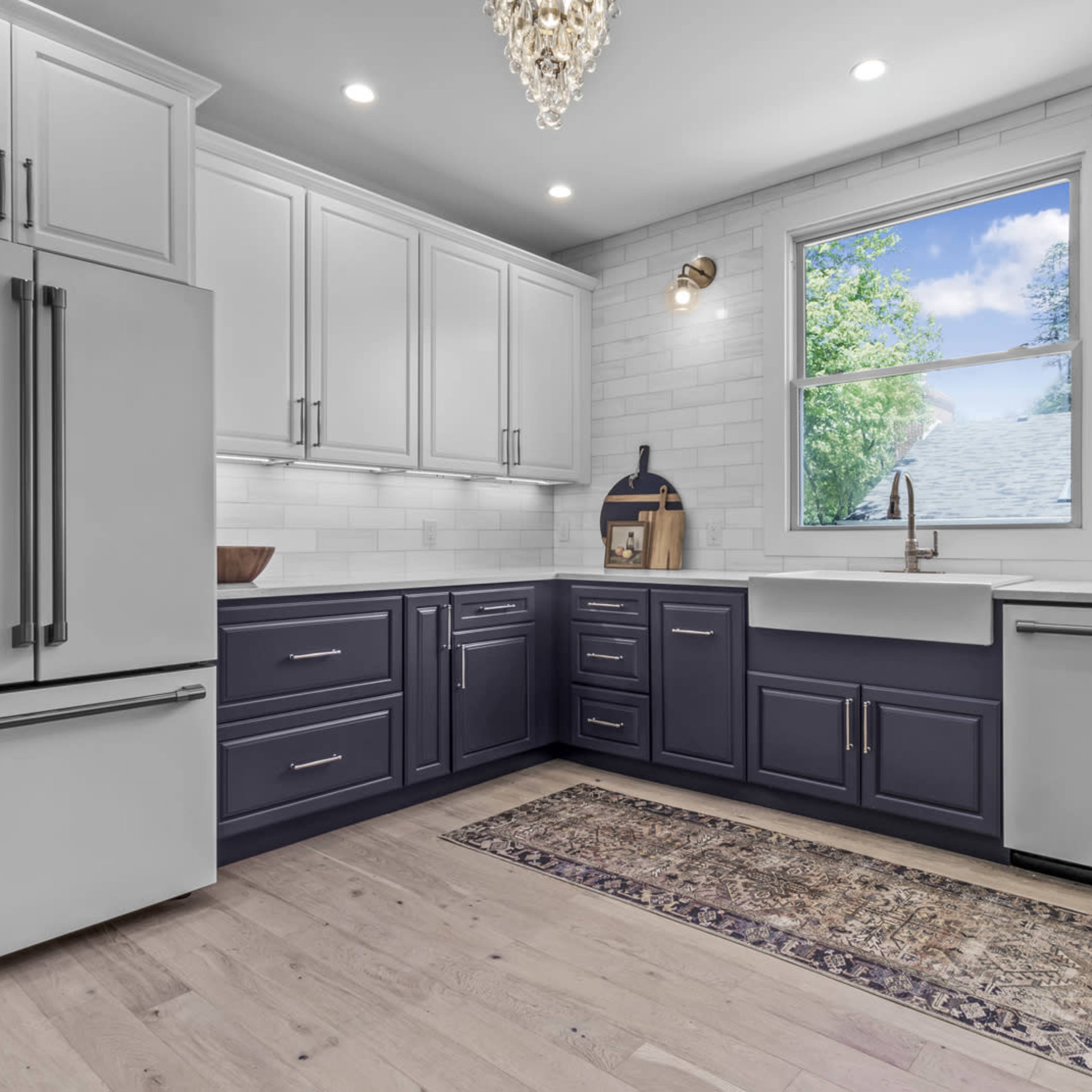 A modern kitchen features white and navy cabinets, a farmhouse sink, and a large window illuminating the space.