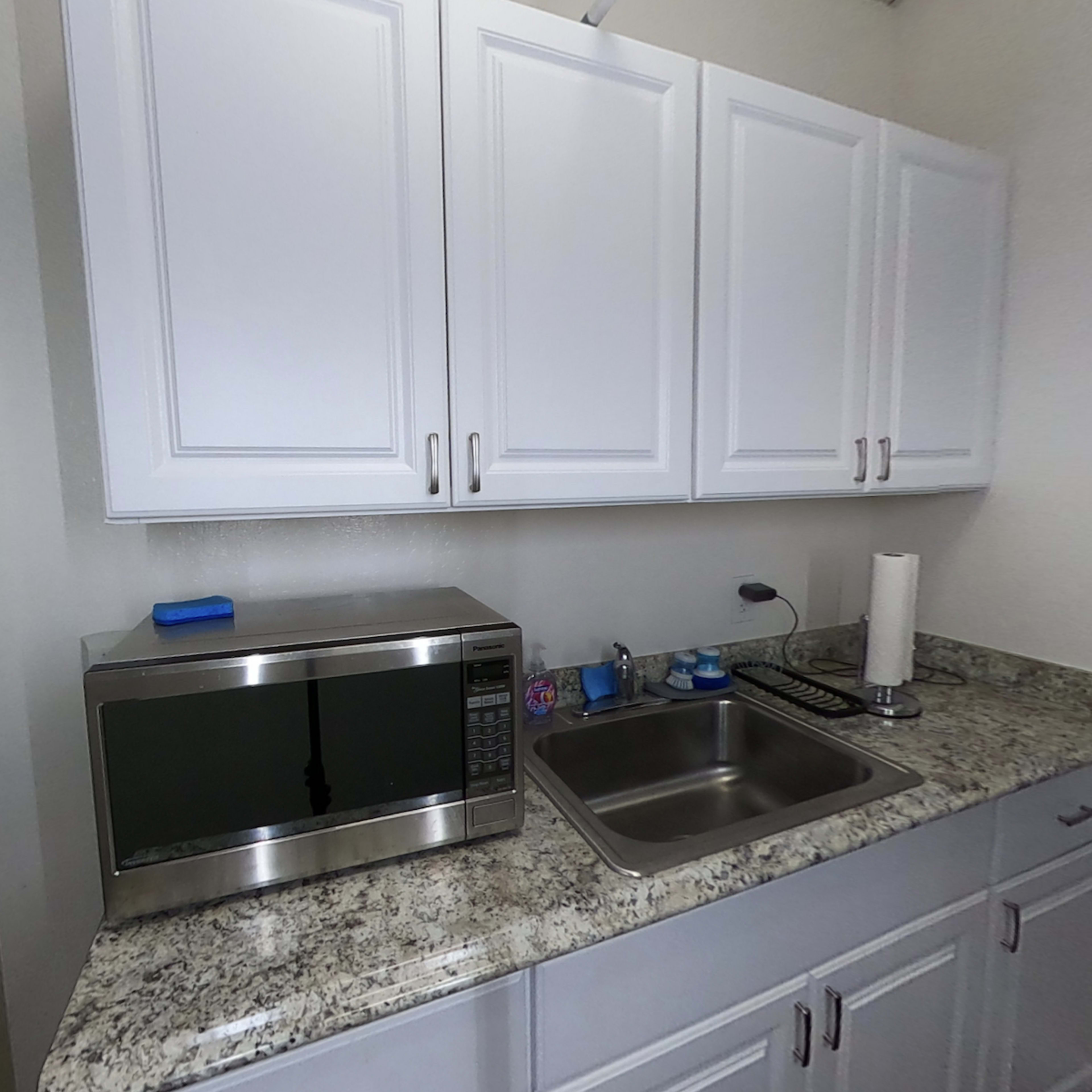 A compact kitchen area features white cabinets, a stainless steel microwave, a sink, and a countertop with various kitchen items.