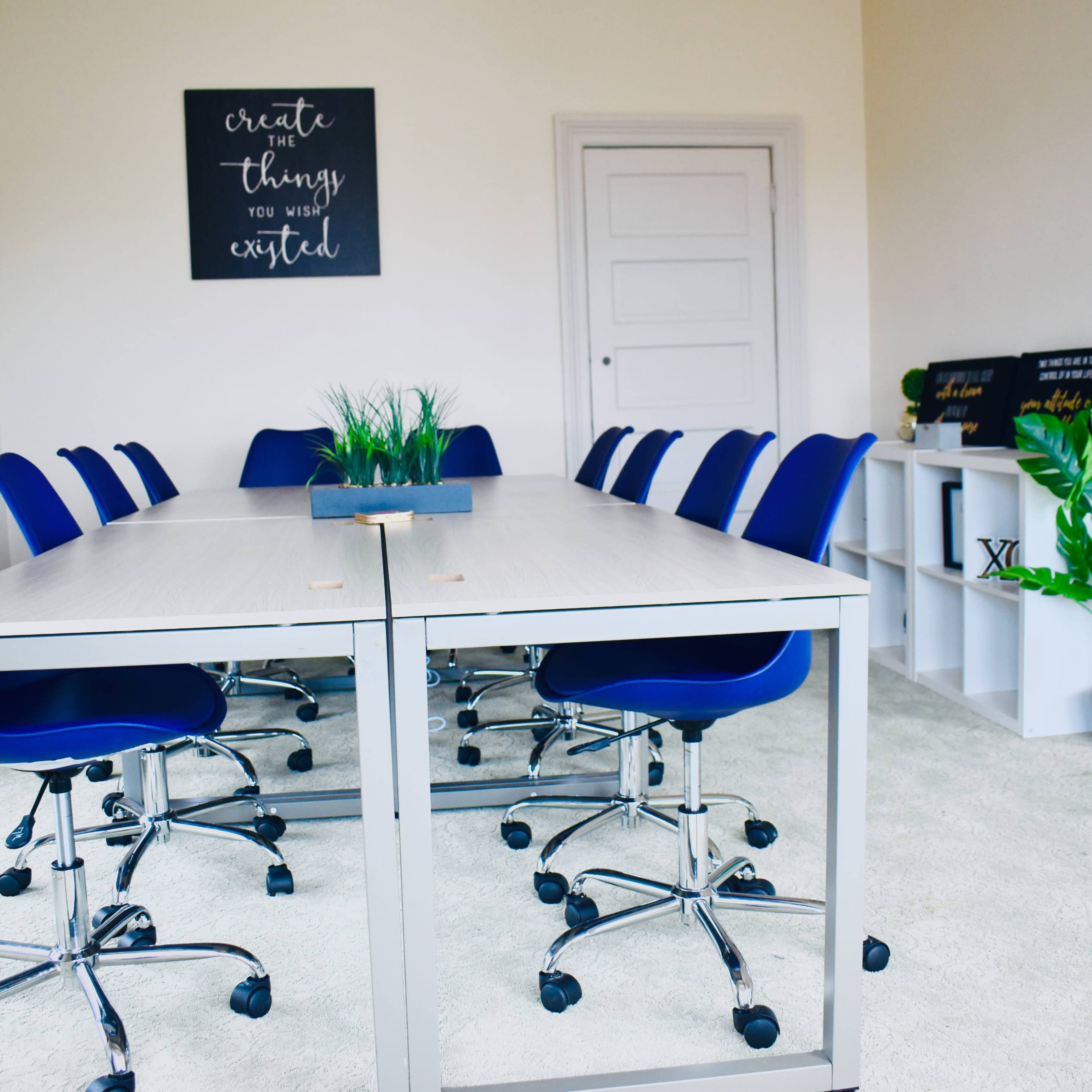 A conference room features a long table surrounded by blue chairs, with greenery and motivational decor on the walls.