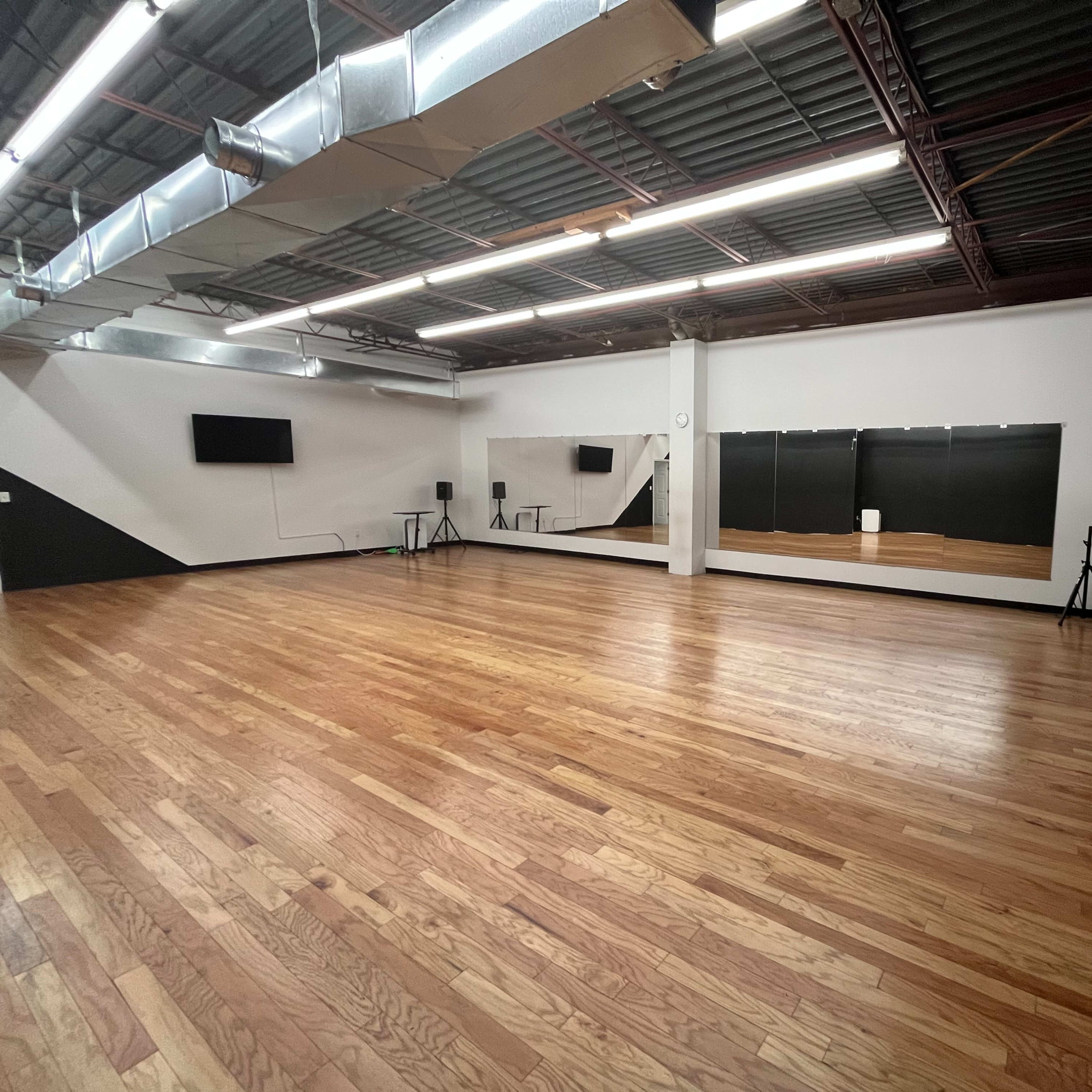 An empty dance studio with wooden floors, mirrors on one wall, and minimal equipment in the corners.