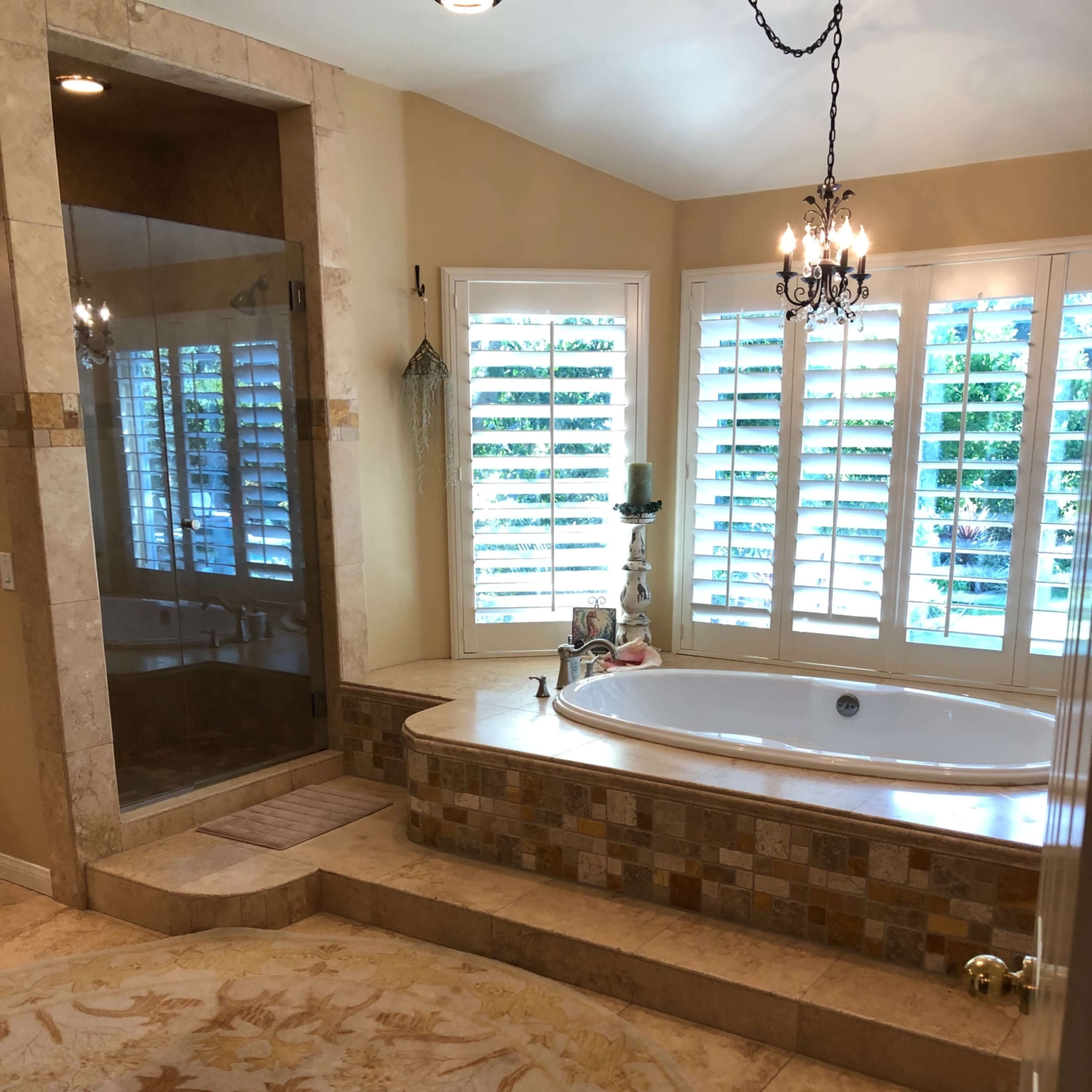 A bathroom featuring a Jacuzzi tub, a glass shower enclosure, and large windows with plantation shutters.