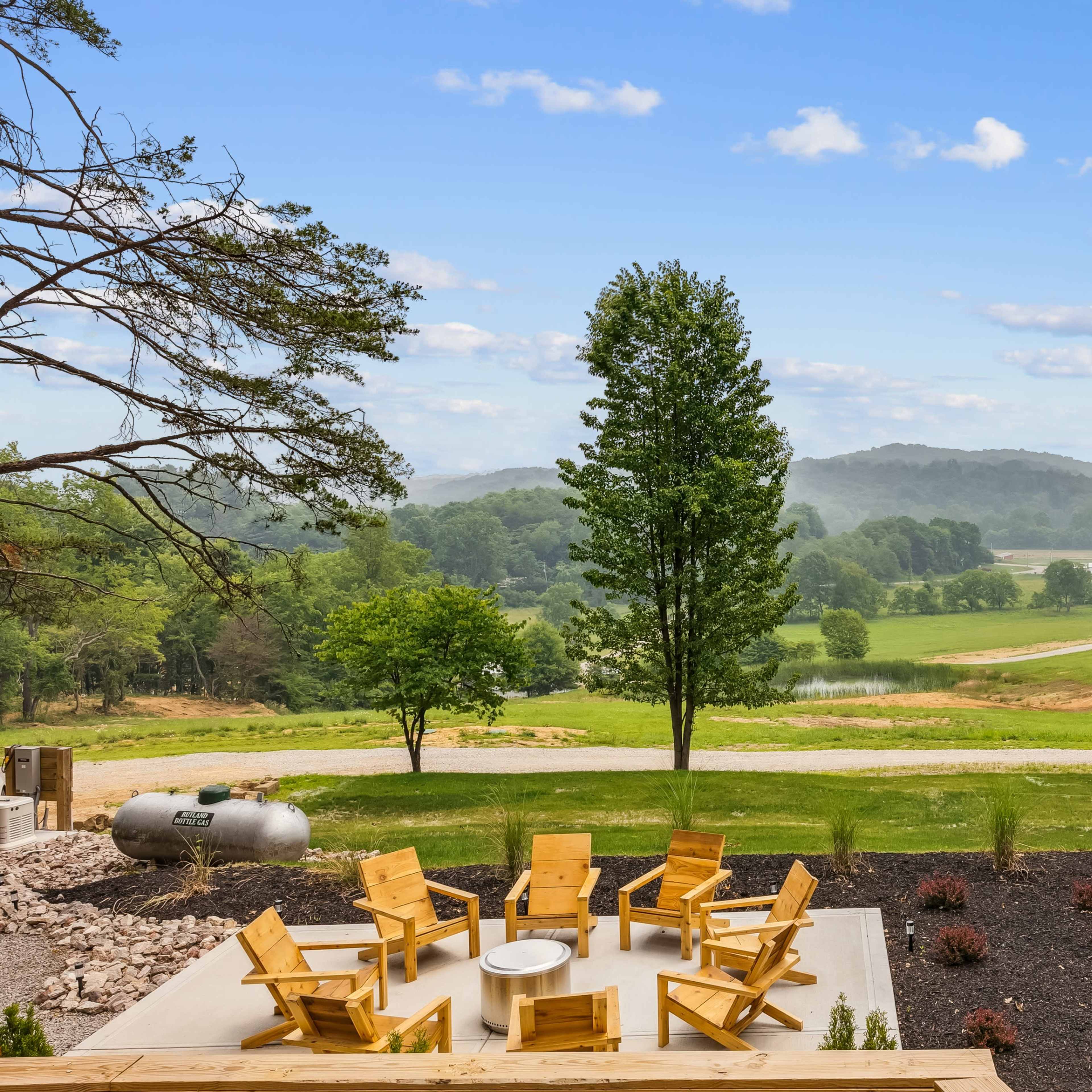 A circular arrangement of wooden chairs surrounds a fire pit, with a lush green landscape and rolling hills in the background.