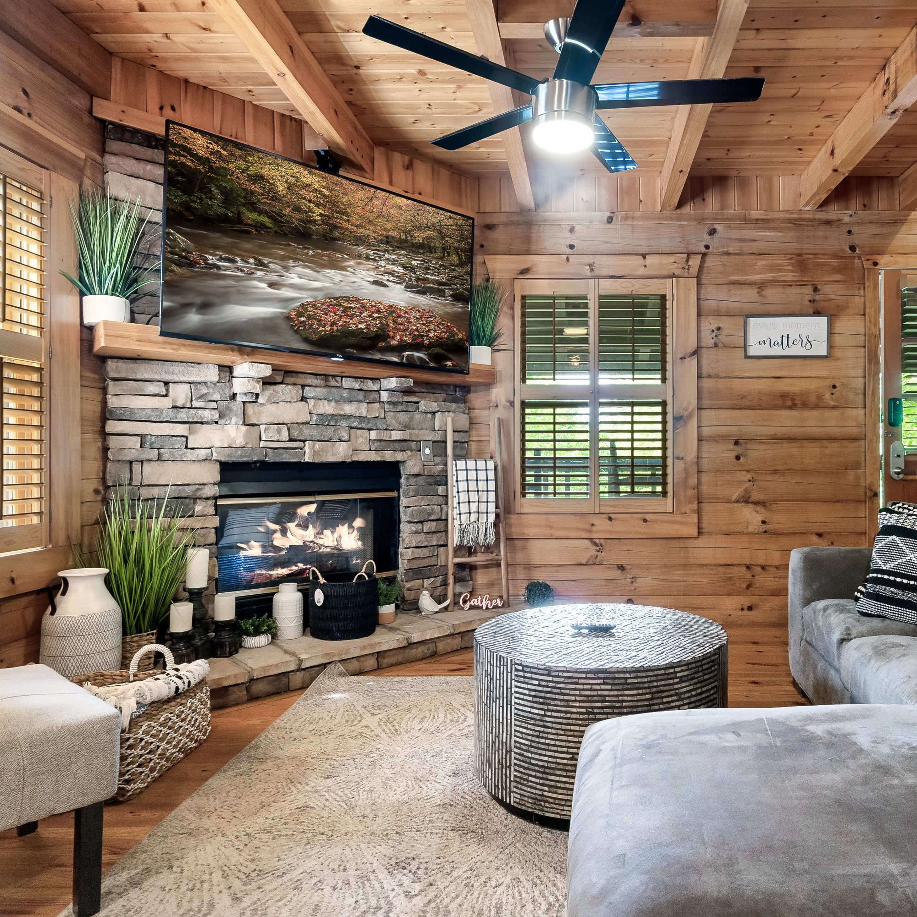 A cozy living room features wooden walls, a stone fireplace, a large television mounted above it, and comfortable seating arranged around a circular table.