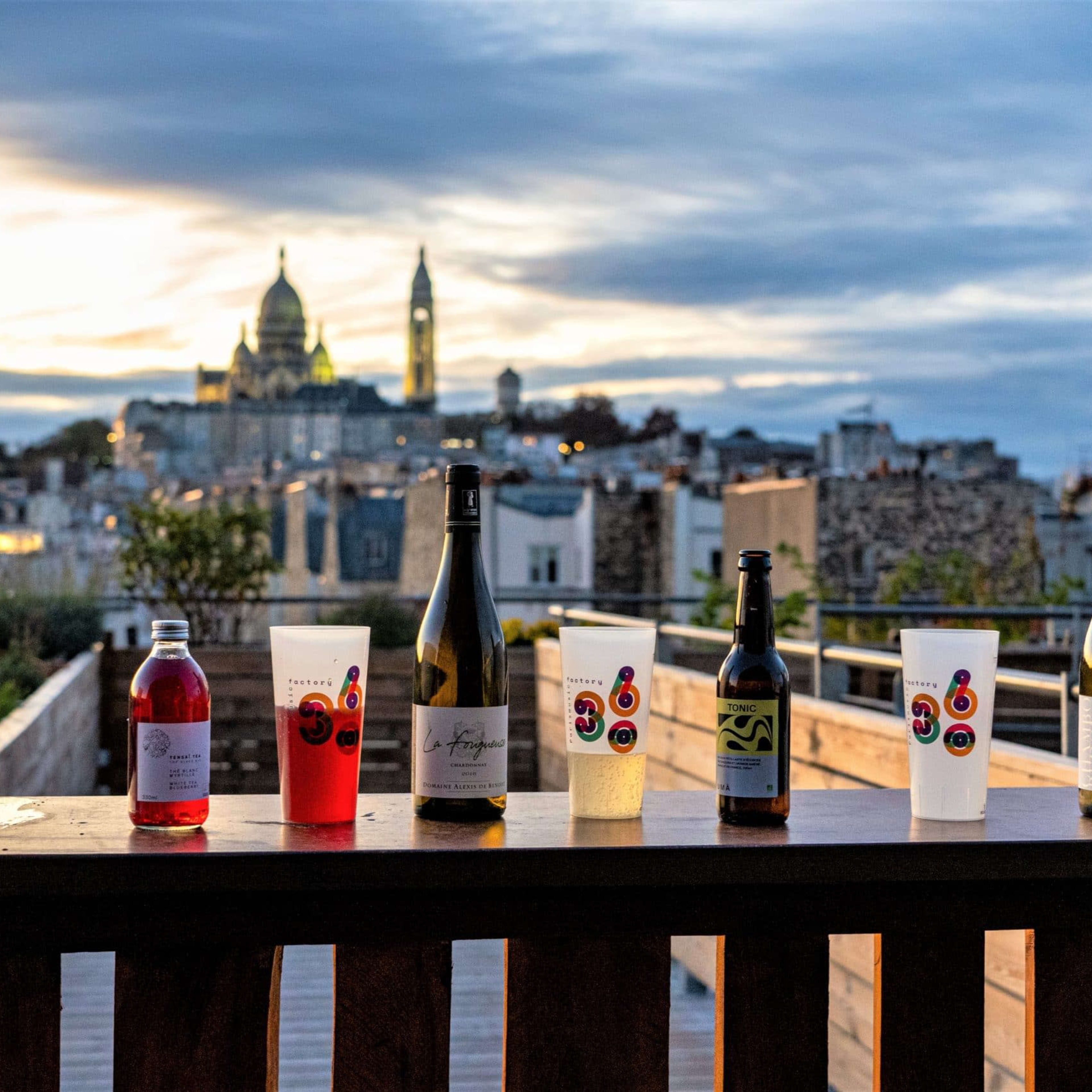 Eight bottles and glasses of various beverages are lined up on a wooden railing with a view of a cityscape at sunset, featuring prominent church domes in the background.
