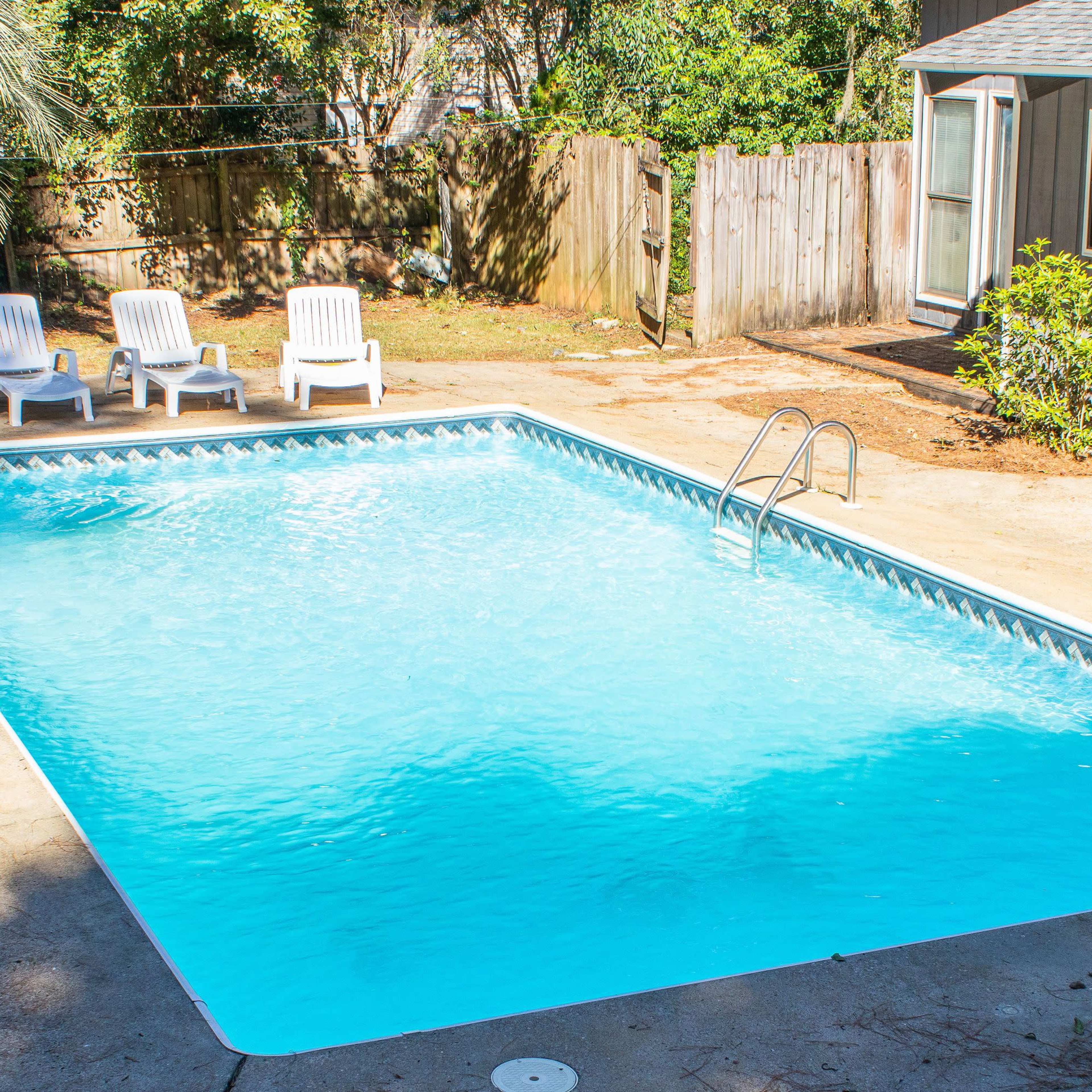 A clear blue swimming pool surrounded by four white lounge chairs and a small gray building in a fenced backyard.