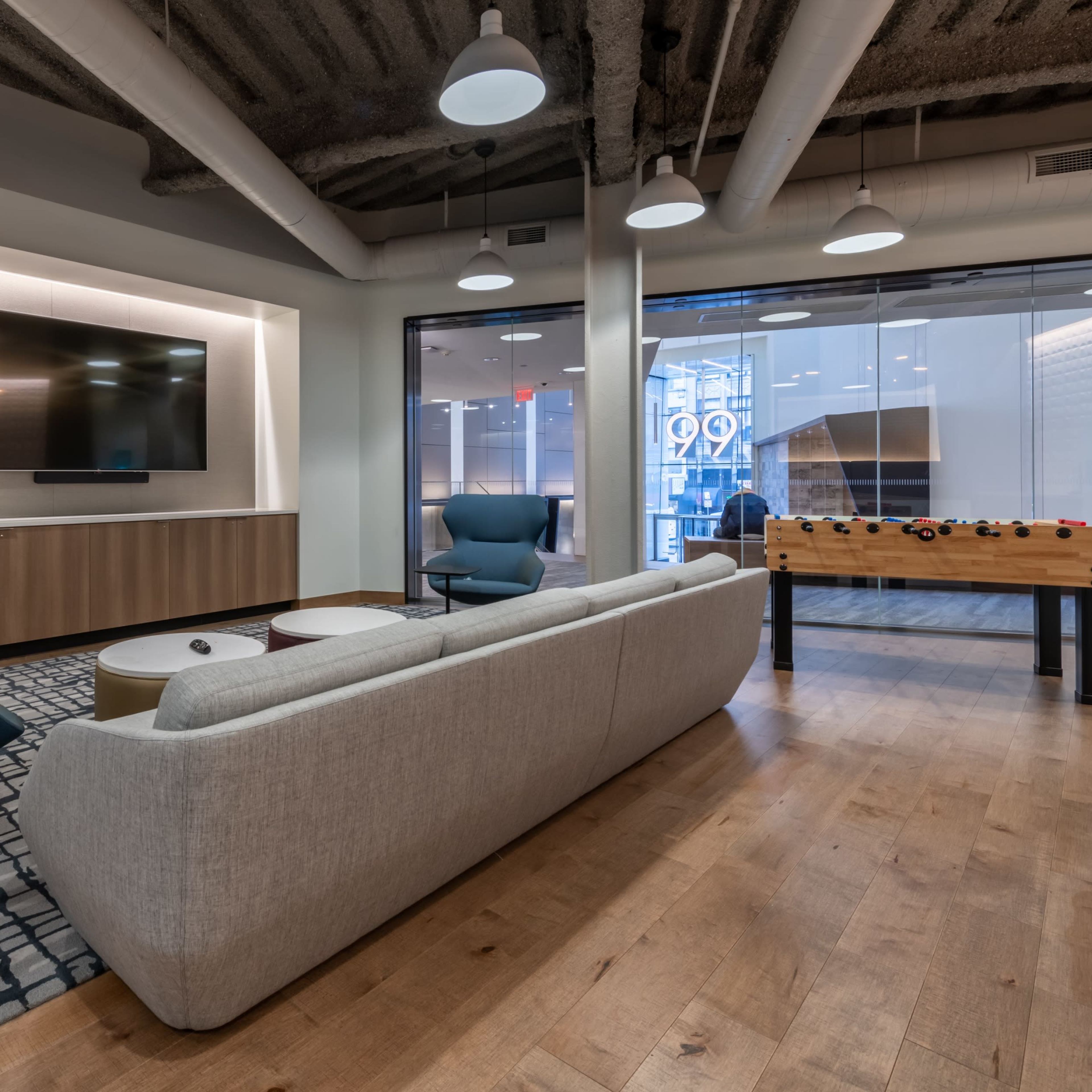 A modern lounge area with a gray sofa, blue chairs, a round coffee table, and a foosball table, all set against a backdrop of large windows.
