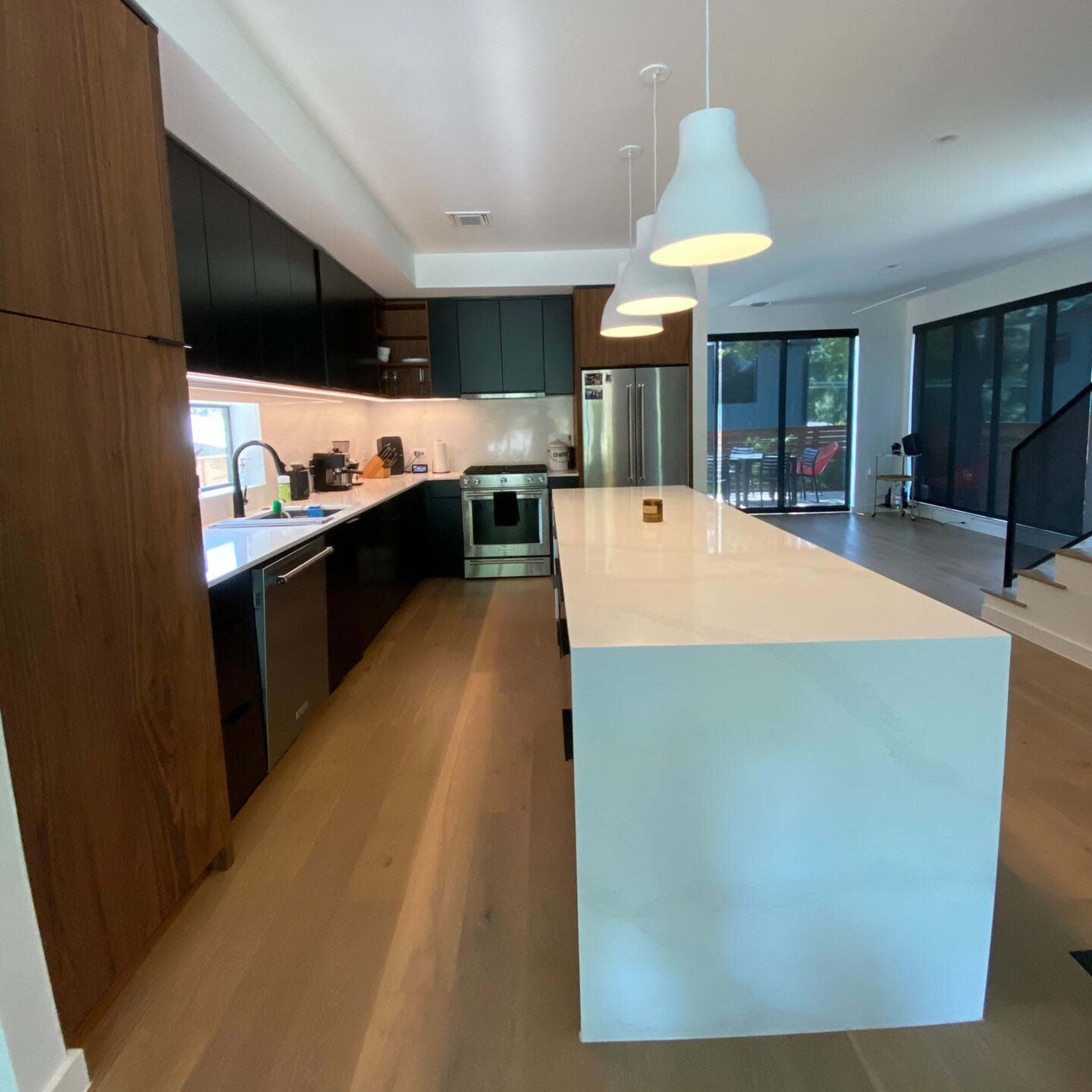 A modern kitchen features dark cabinetry, a white countertop island with pendant lighting, and large windows offering natural light.