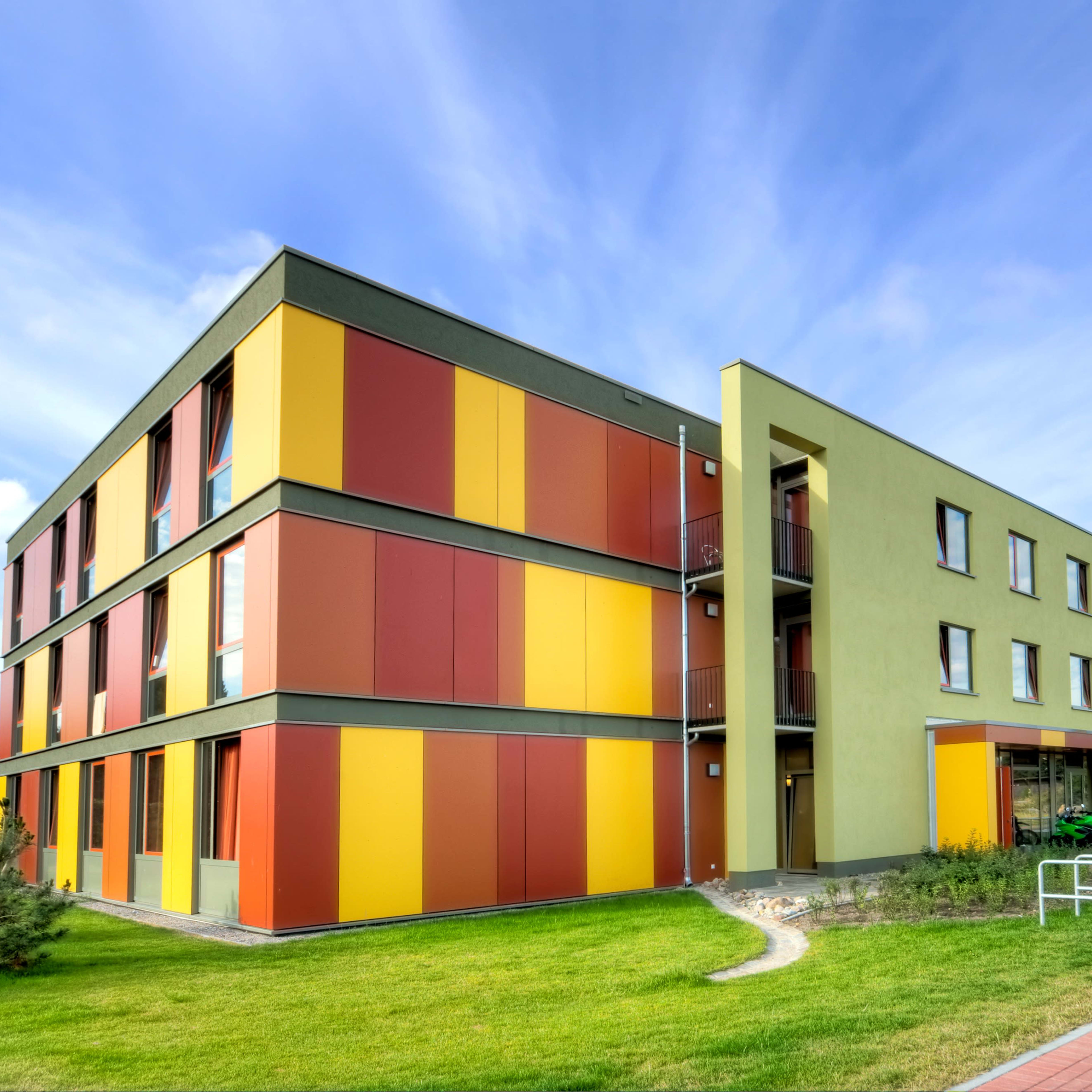 A three-story building features a modern design with colorful yellow and red paneling, surrounded by a grassy area and a bicycle rack.