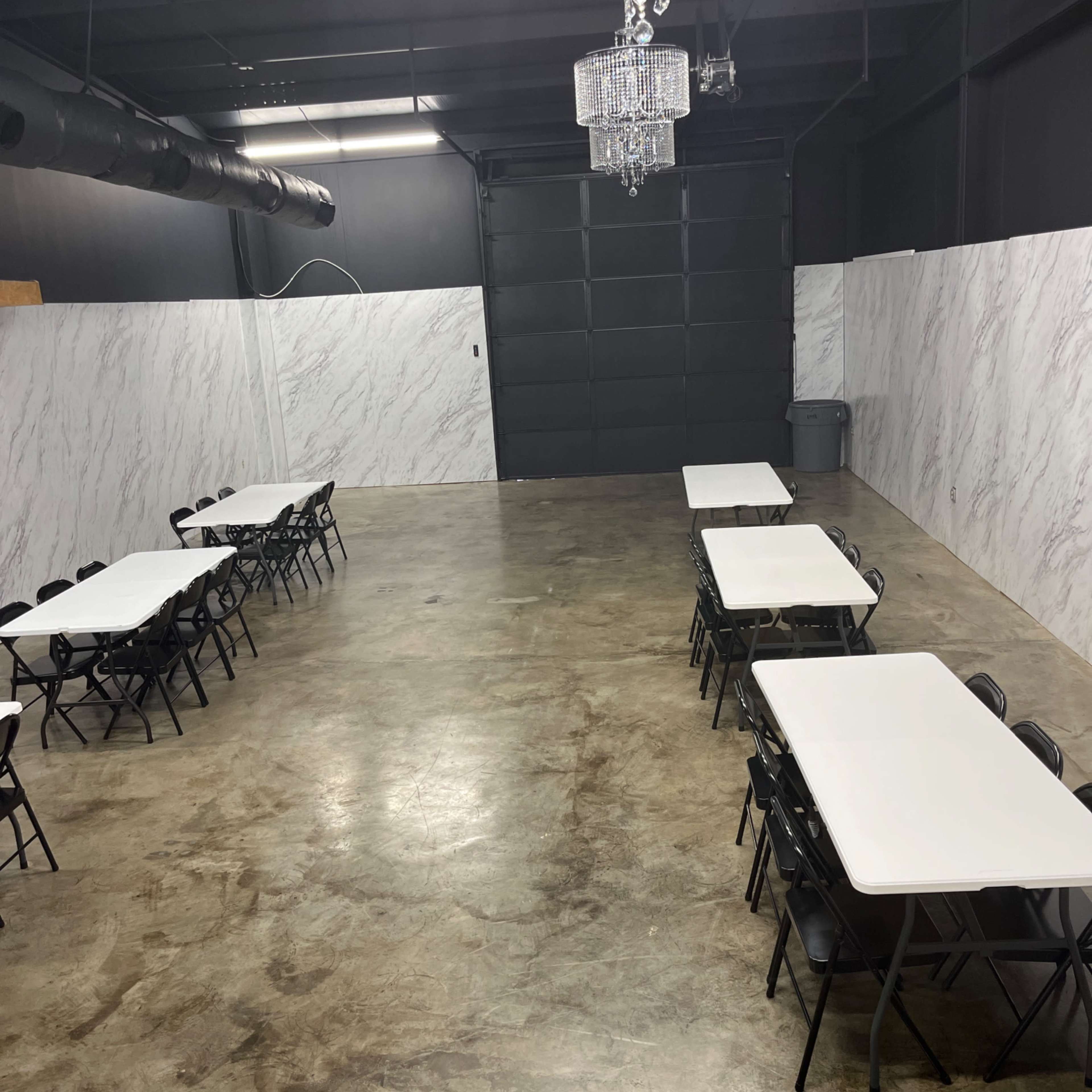 A spacious room with polished concrete floors, featuring several rows of white tables and black chairs, and two chandeliers hanging from the ceiling.