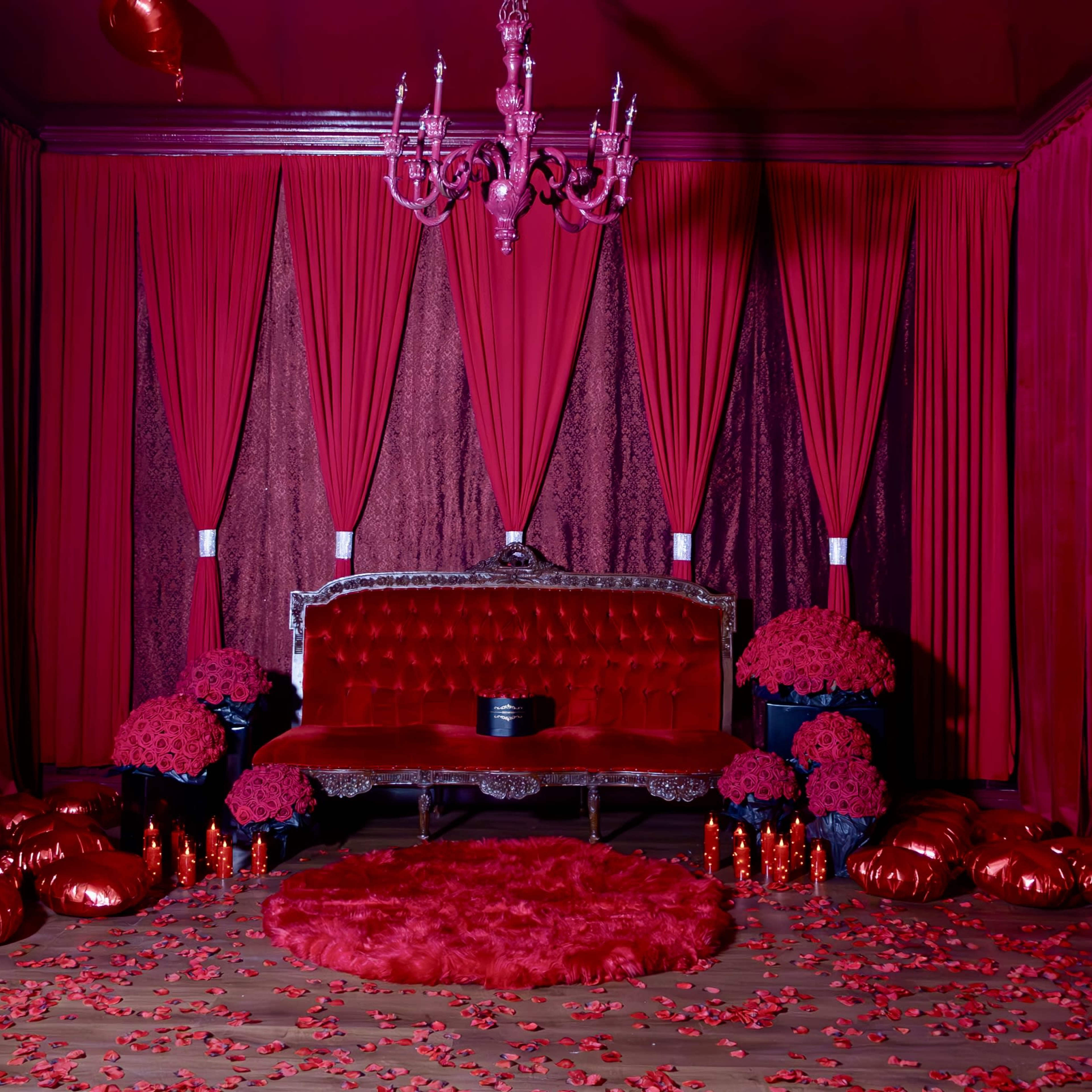 The scene features a richly decorated room with red walls, a velvet sofa, and an array of red decorations, including flowers and pillows scattered across the floor.