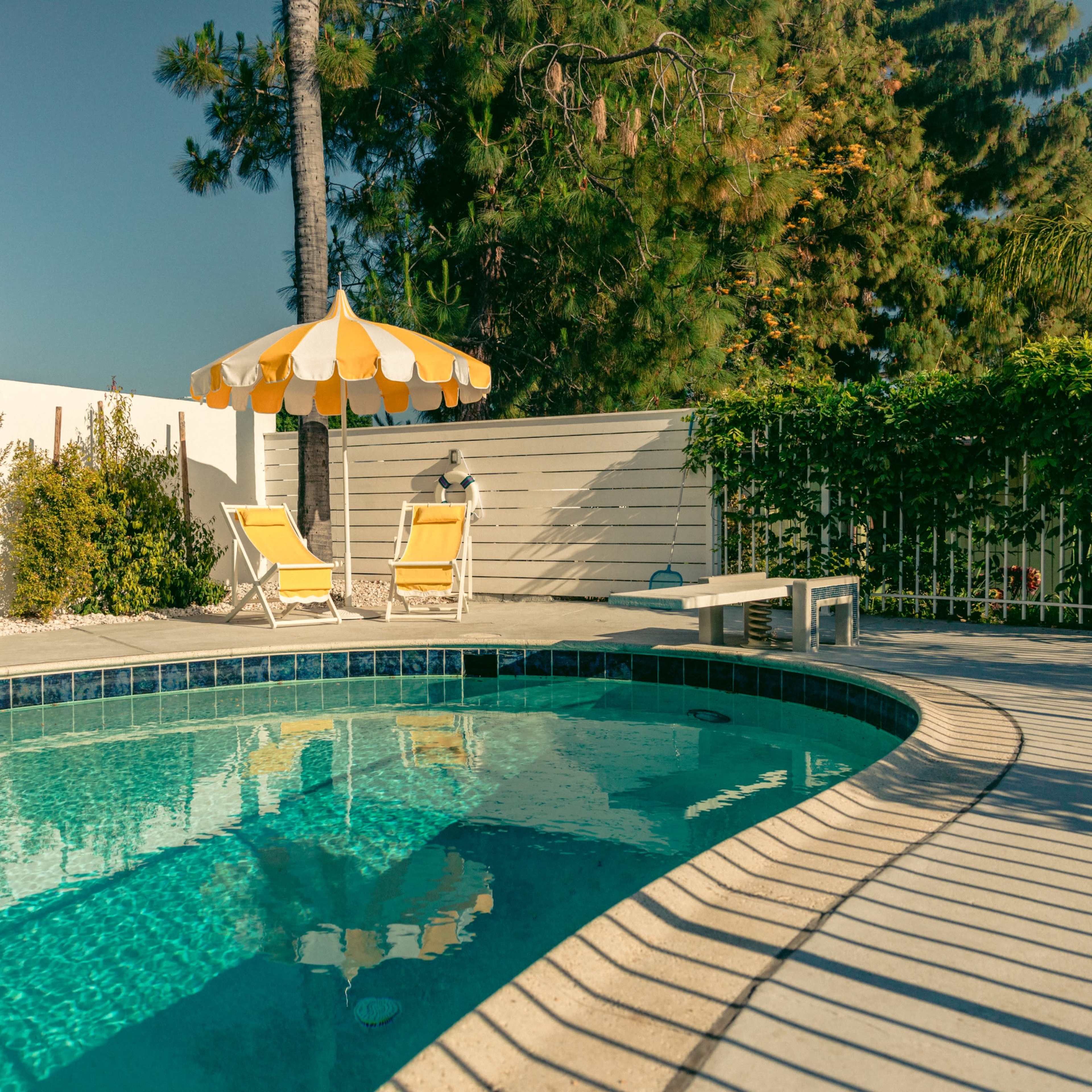 A clear swimming pool is surrounded by lounge chairs and a sun umbrella, with greenery and a white fence in the background.