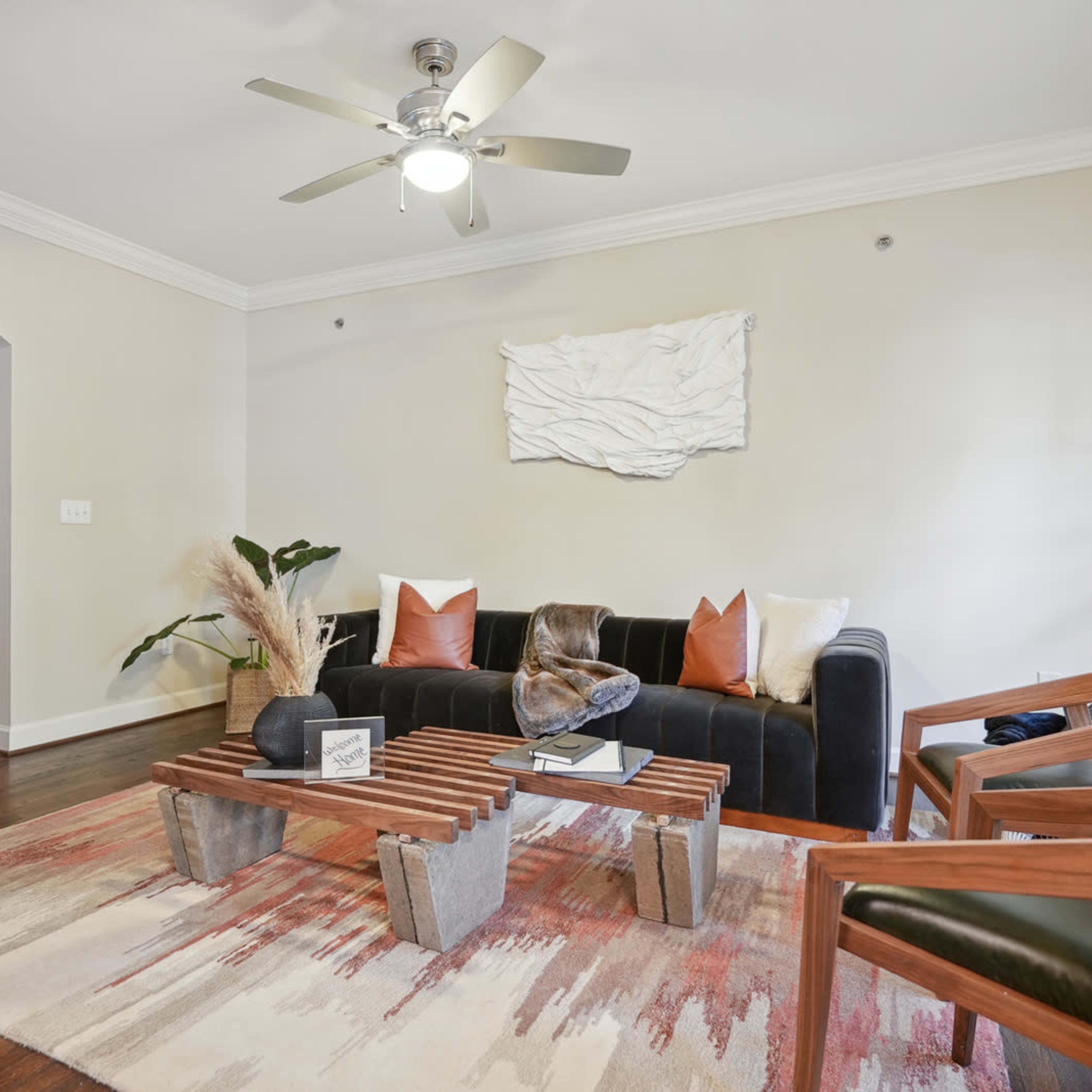 The living room features a black sofa, a wooden coffee table, and two armchairs arranged on a patterned rug, with a ceiling fan above and a decorative wall piece.