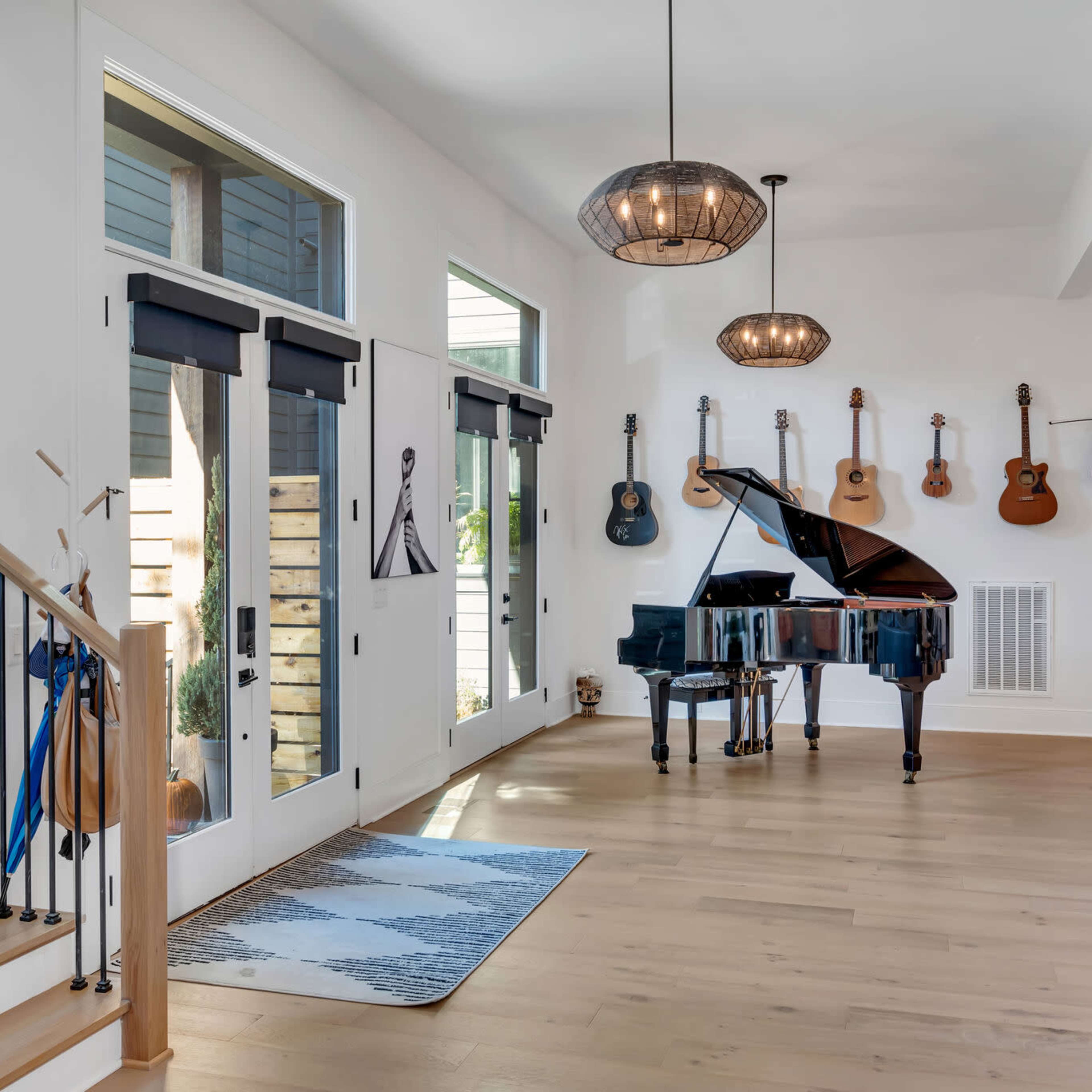 A spacious living area featuring a black grand piano, several hanging guitars on the wall, and large windows allowing natural light to enter.