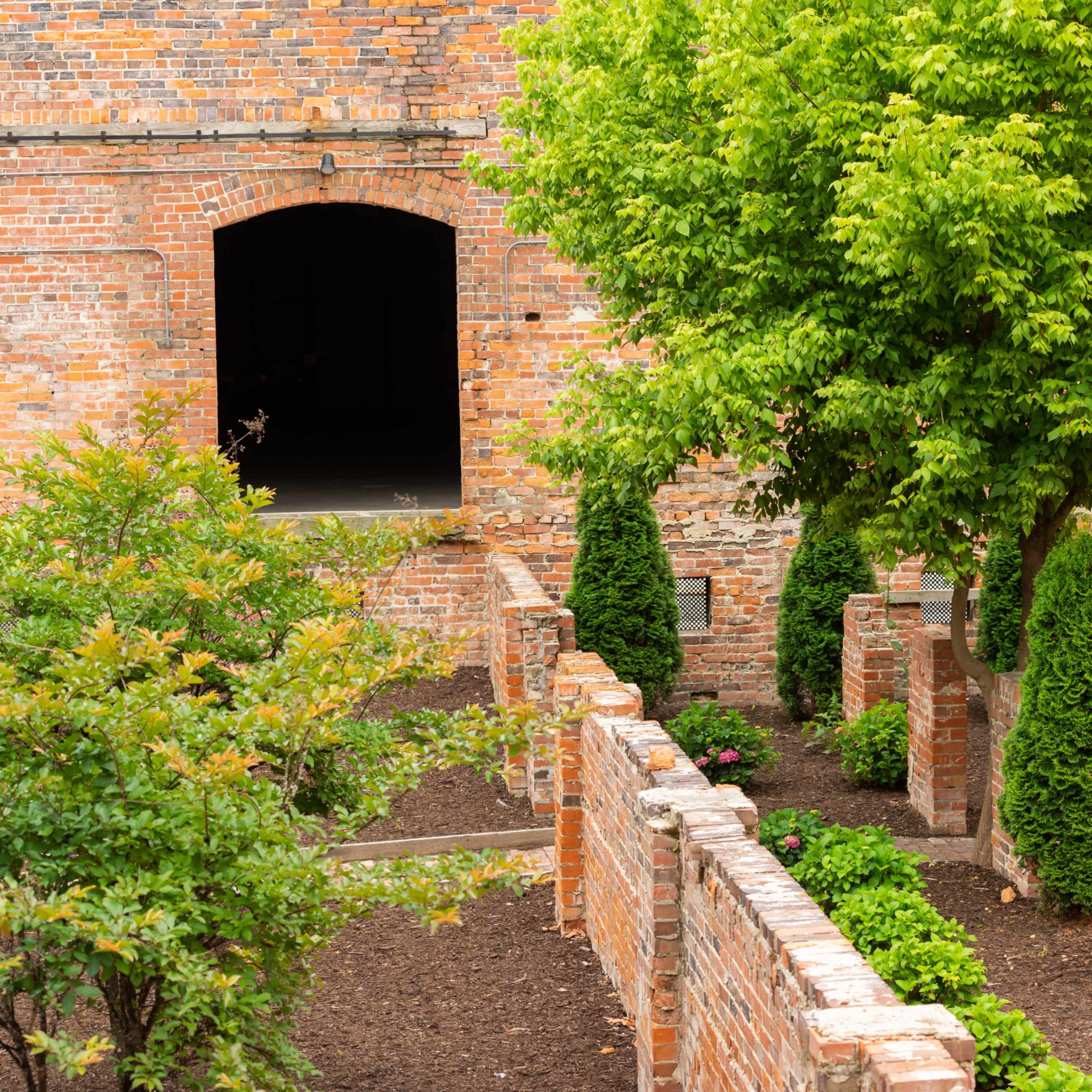 A green garden with neatly trimmed shrubs and a tree is surrounded by a brick wall, and a large archway leads to a dark interior space.
