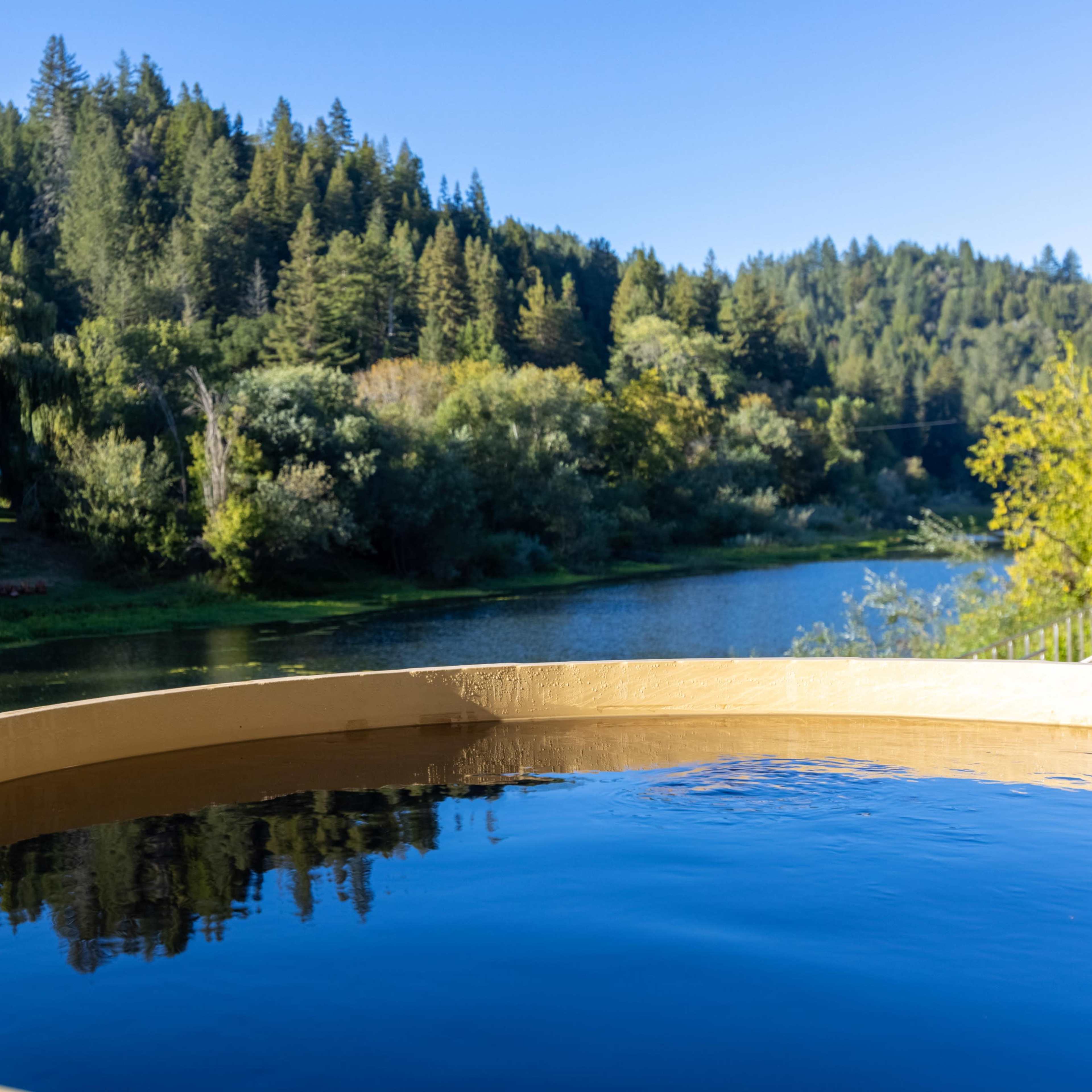 A circular wooden hot tub sits on the edge of a serene lake surrounded by lush green trees and a clear blue sky.