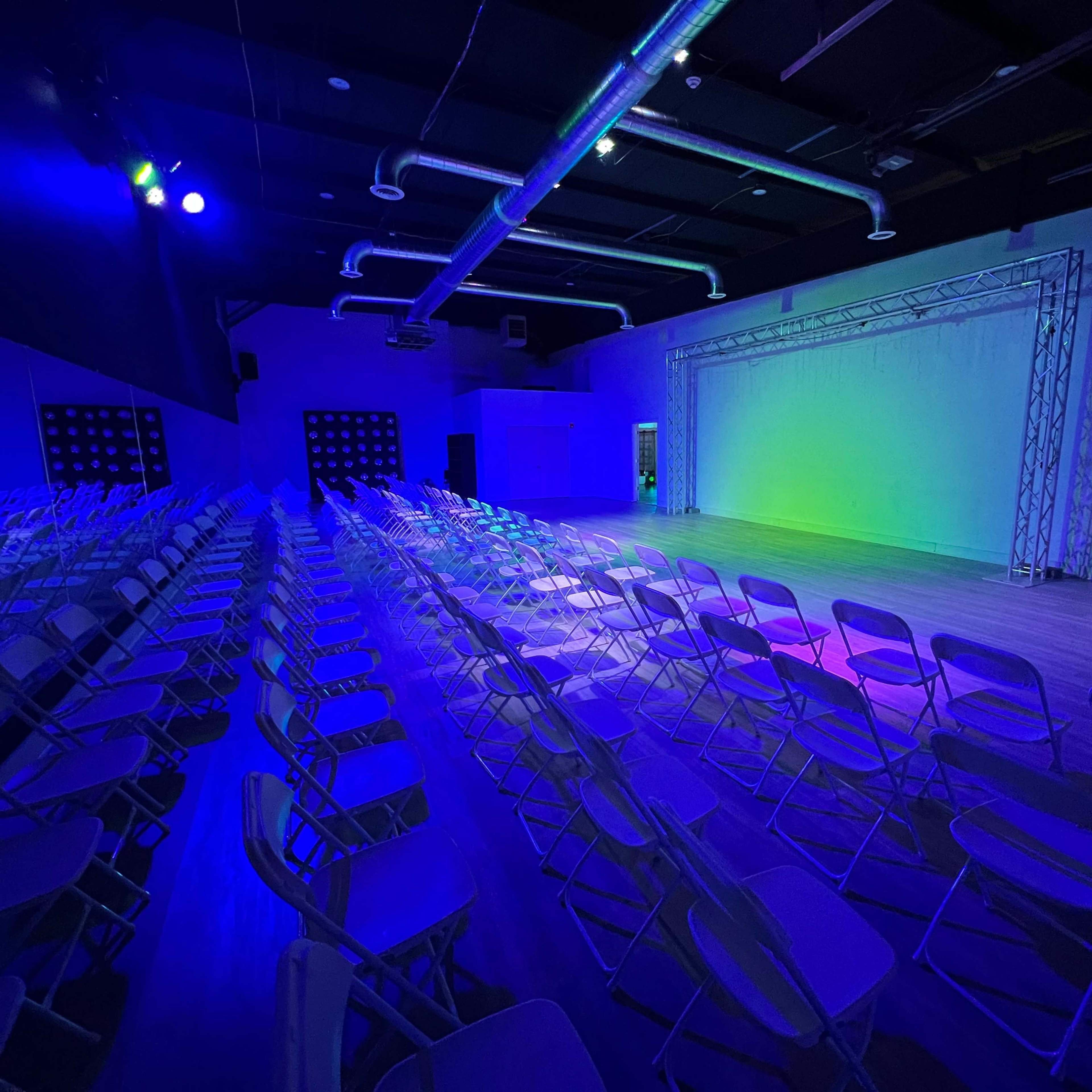A large indoor space with rows of white chairs arranged in front of a simple stage backdrop, illuminated by blue and green lights.